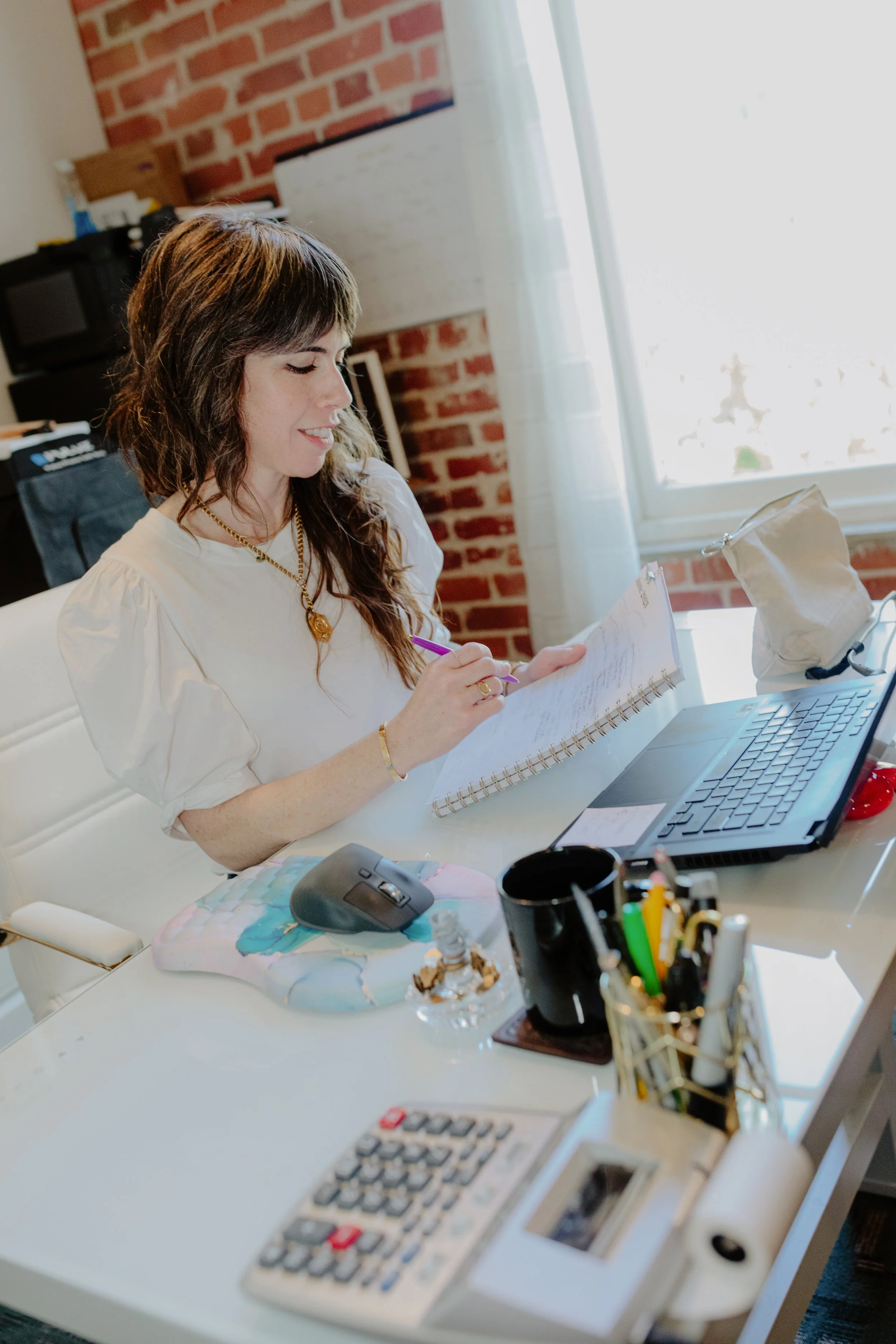 A woman with shoulder-length brown hair and wearing a white blouse is sitting at a desk, writing in a notebook with a purple pen. The desk has a computer mouse, a calculator, and a display of pens and markers. There is a window with sheer curtains be
