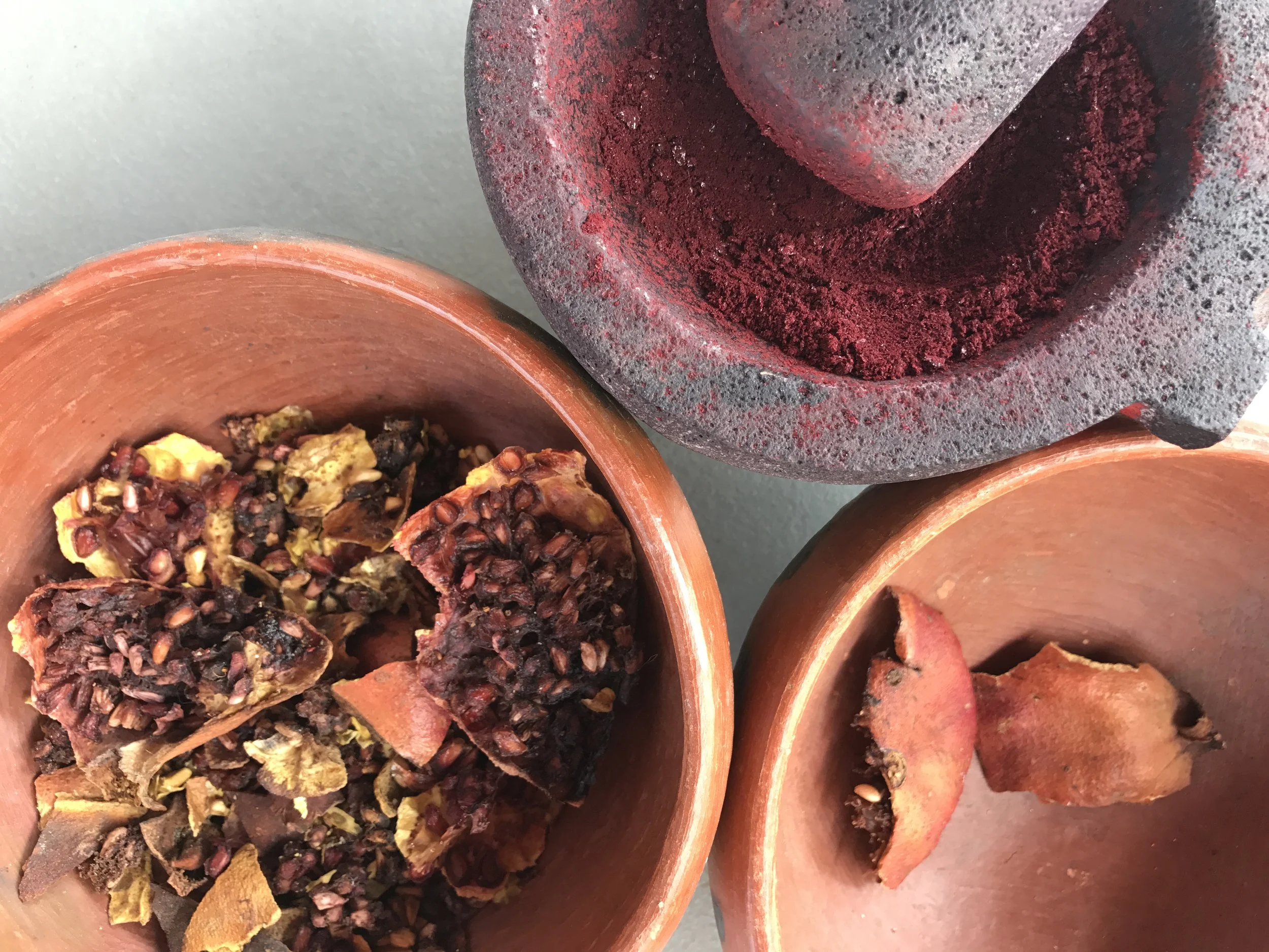 Close-up of a mortar and pestle with red herbal powder. Part of a clay pot with chopped herbs or dried flowers visible at the bottom.