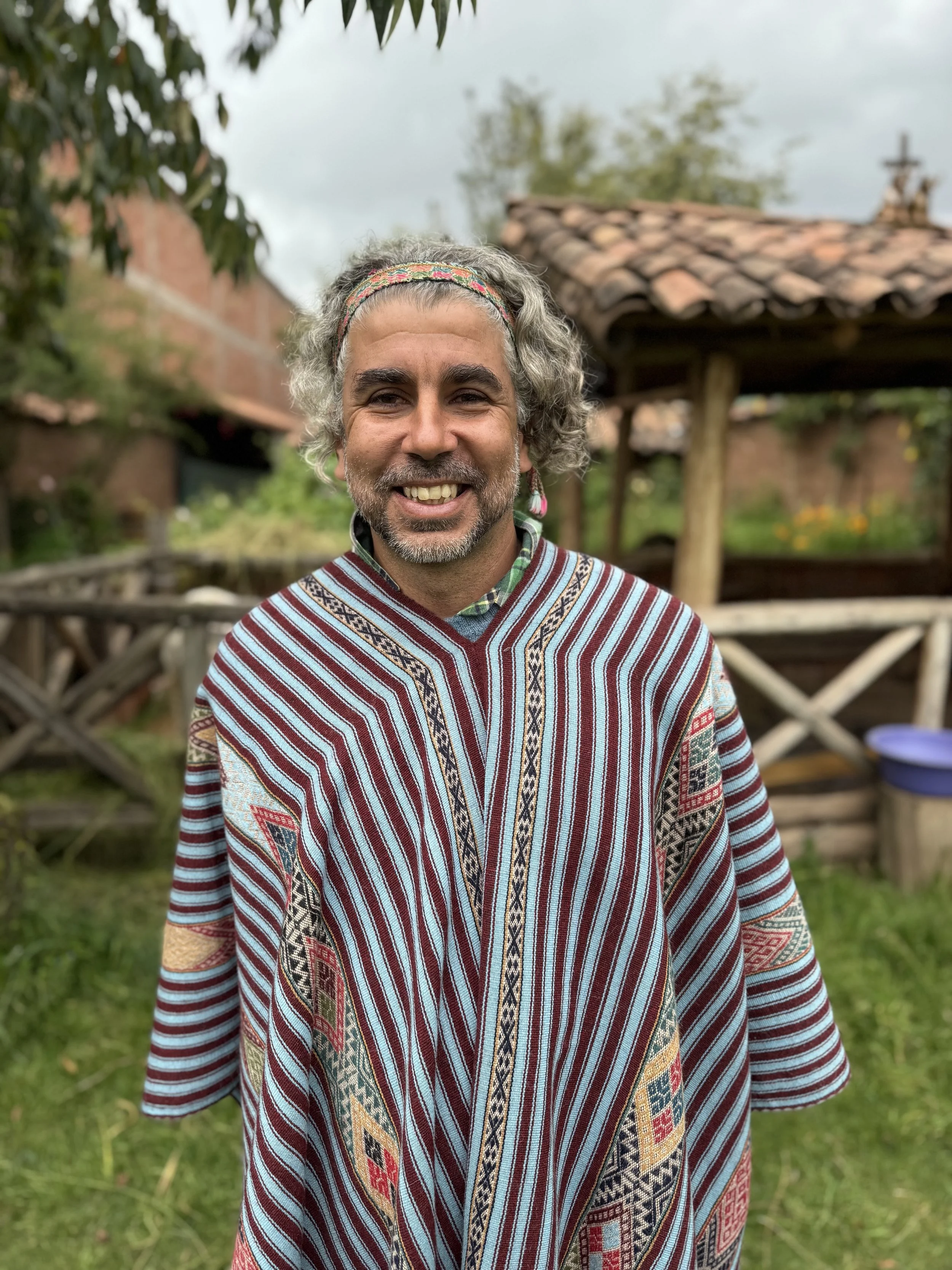 A smiling man with gray curly hair and a beard, wearing a colorful striped traditional garment, standing outdoors with a wooden fence, greenery, and a building with a tiled roof in the background.