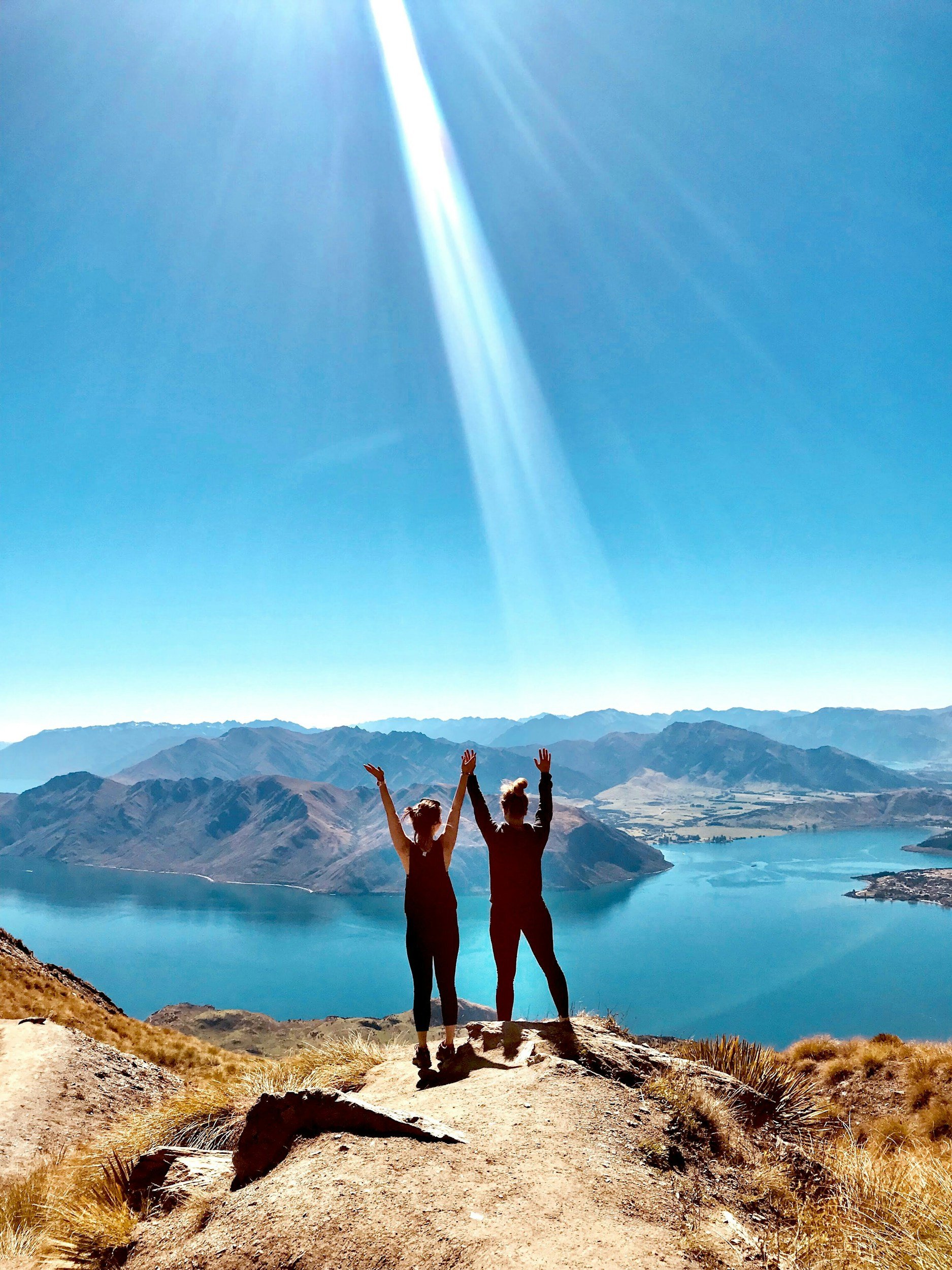 Two women standing on a rocky hilltop with arms raised, overlooking a large blue lake surrounded by mountain ranges under a bright blue sky with sunlight.