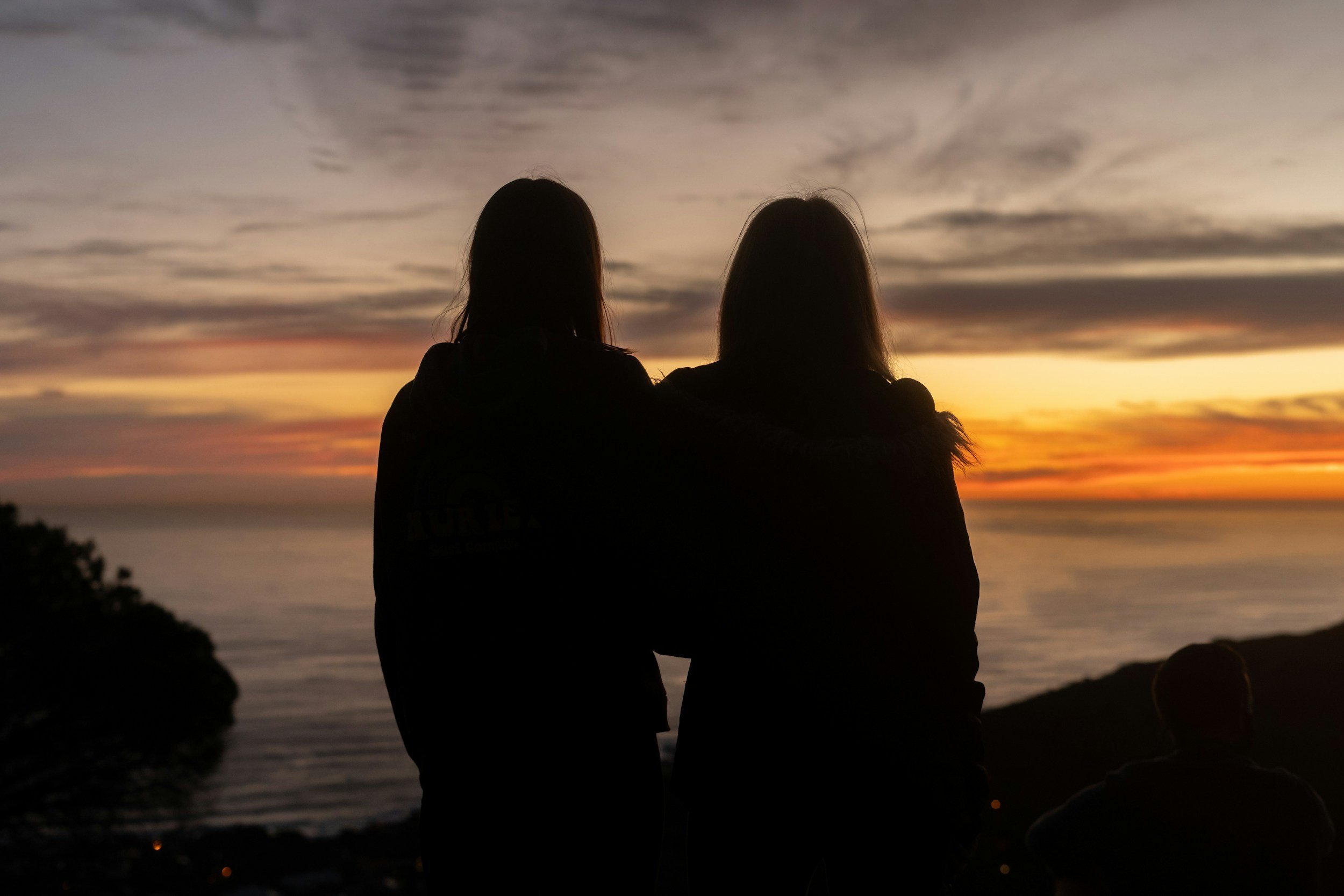 Silhouette of two women sitting side by side, watching a sunset over the ocean with a cloudy sky.