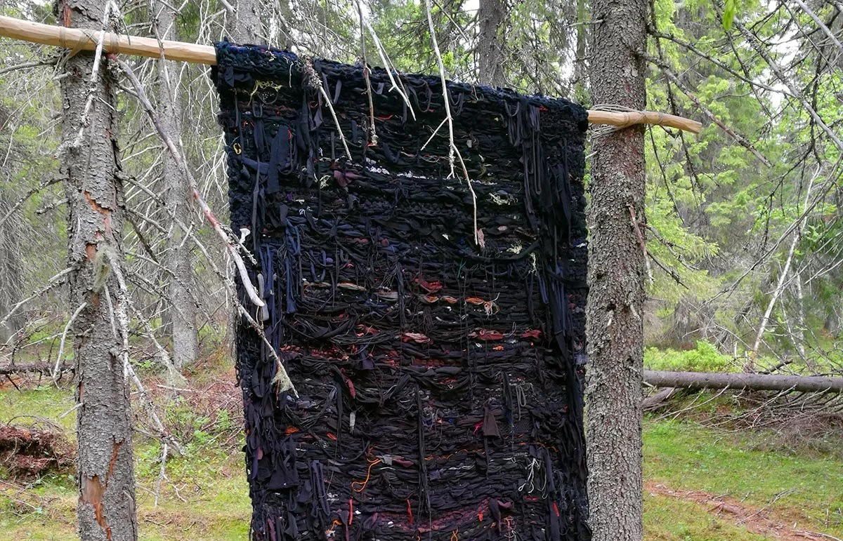 Vainion räsymattoa muistuttava, pääsävyltään musta tekstiiliteos roikkuu riu’ulla kahden ohuen kuusen välissä.