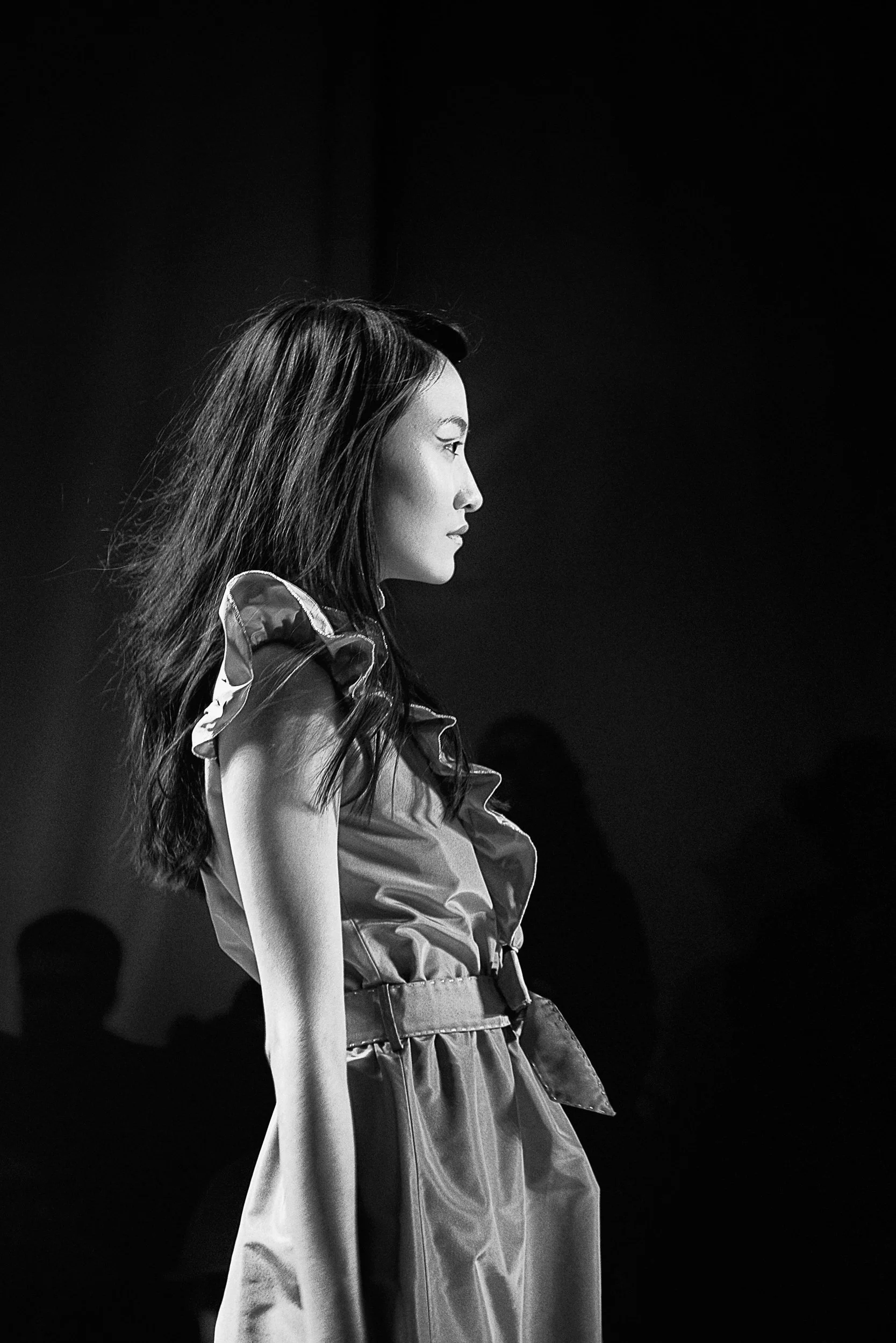 Black and white photo of a woman with long hair, dressed in a silky dress with puffy sleeves, standing in profile against a dark background.