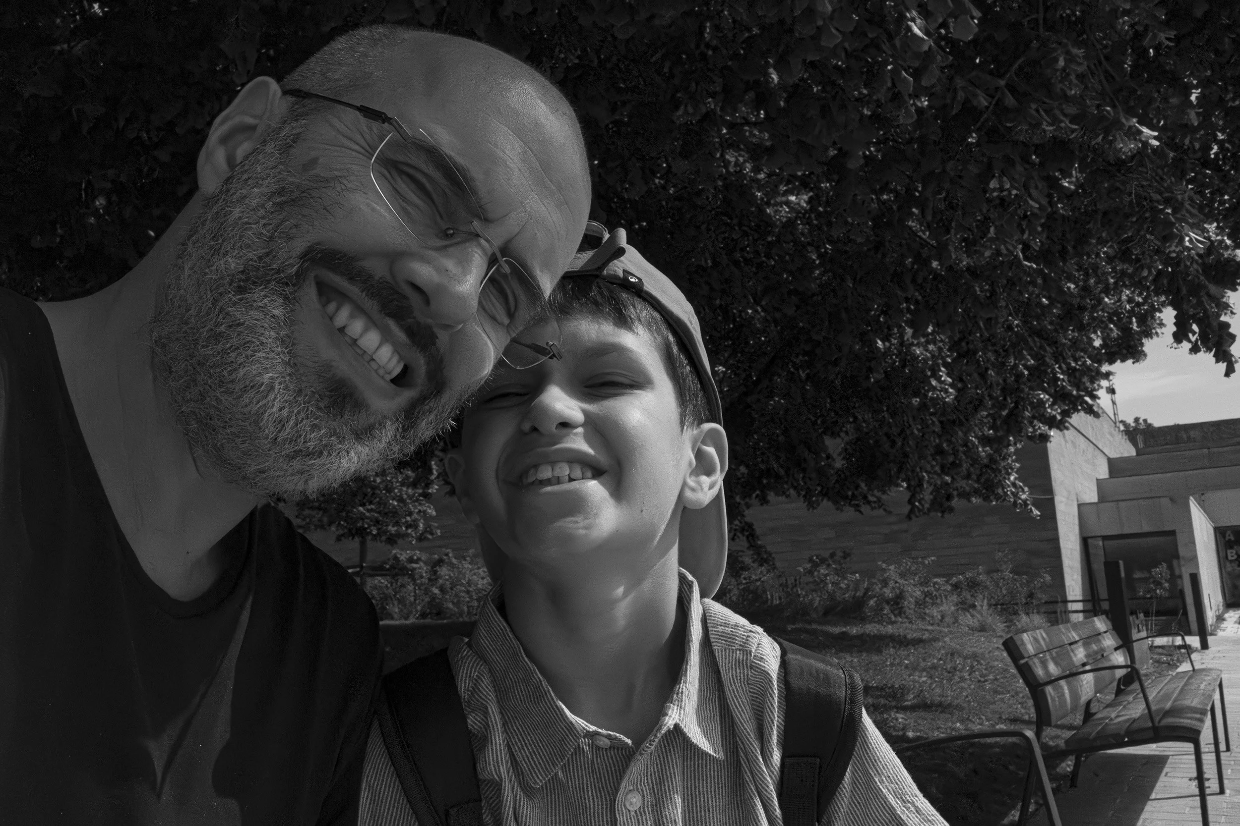 A man and a young boy smiling and taking a selfie outdoors, with trees and a building in the background.