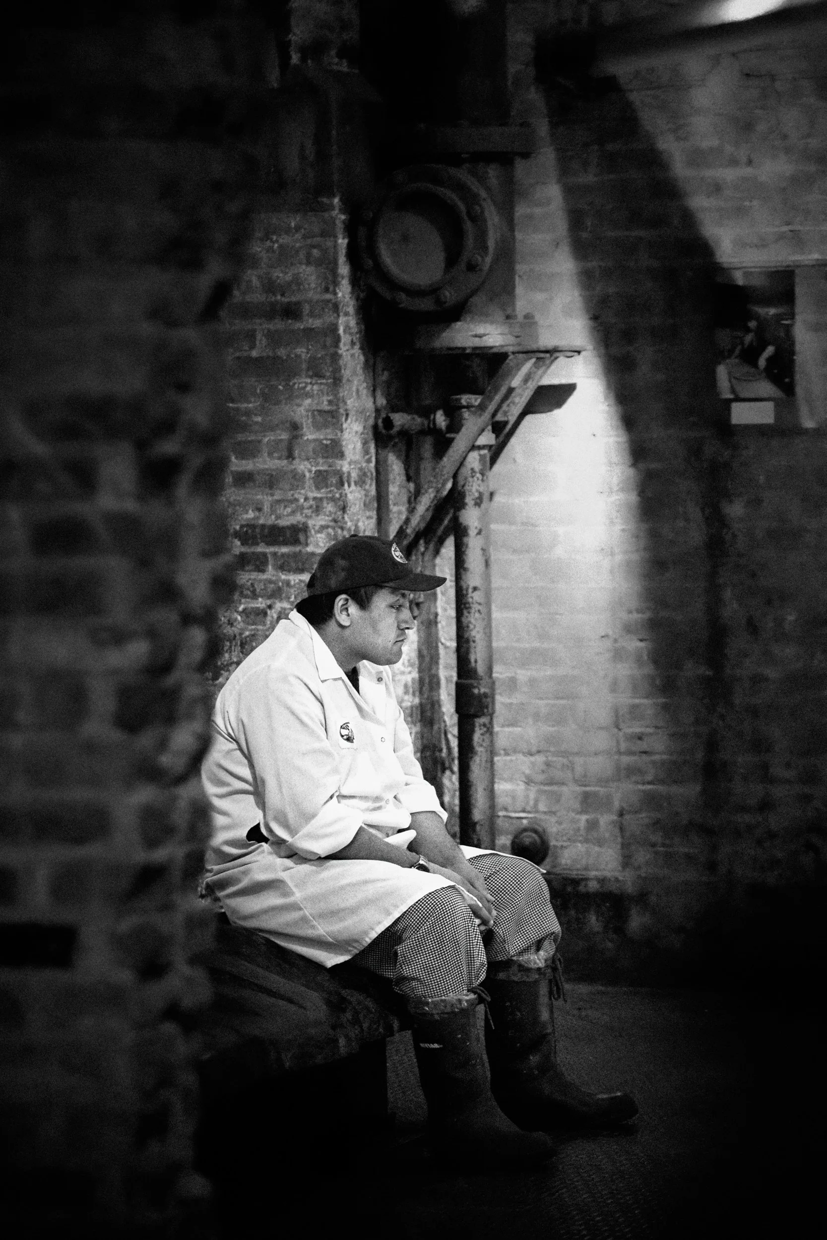 A young person in a chef uniform sits against a brick wall in a dimly lit room, with elbows resting on knees, looking contemplatively downward.