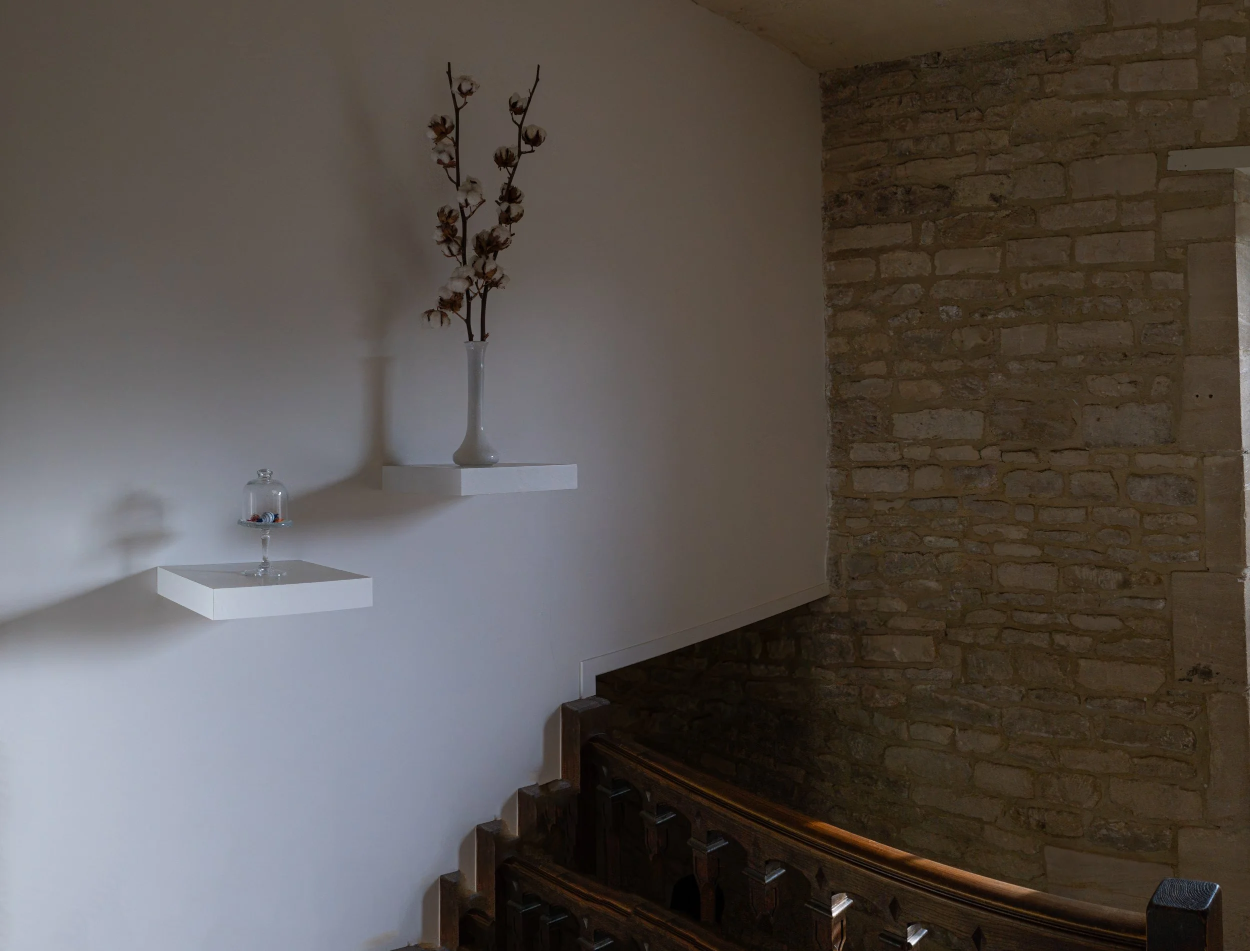 A minimalistic interior with a white shelf holding a glass vase with cotton stems and a small glass dome with decorative objects, positioned against a plain white wall and an adjacent exposed brick wall.