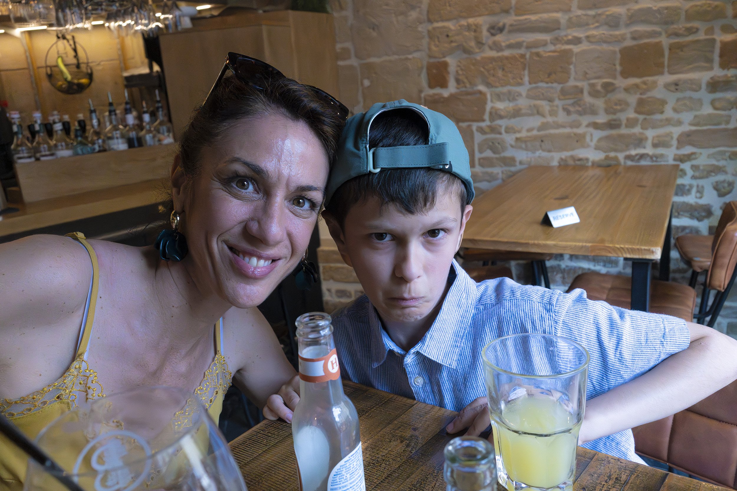 A woman and a young boy taking a selfie at a restaurant. The woman is smiling with sunglasses on her head, wearing a yellow dress with floral details. The boy is wearing a blue cap backward and a striped shirt, with a serious expression. They are sit