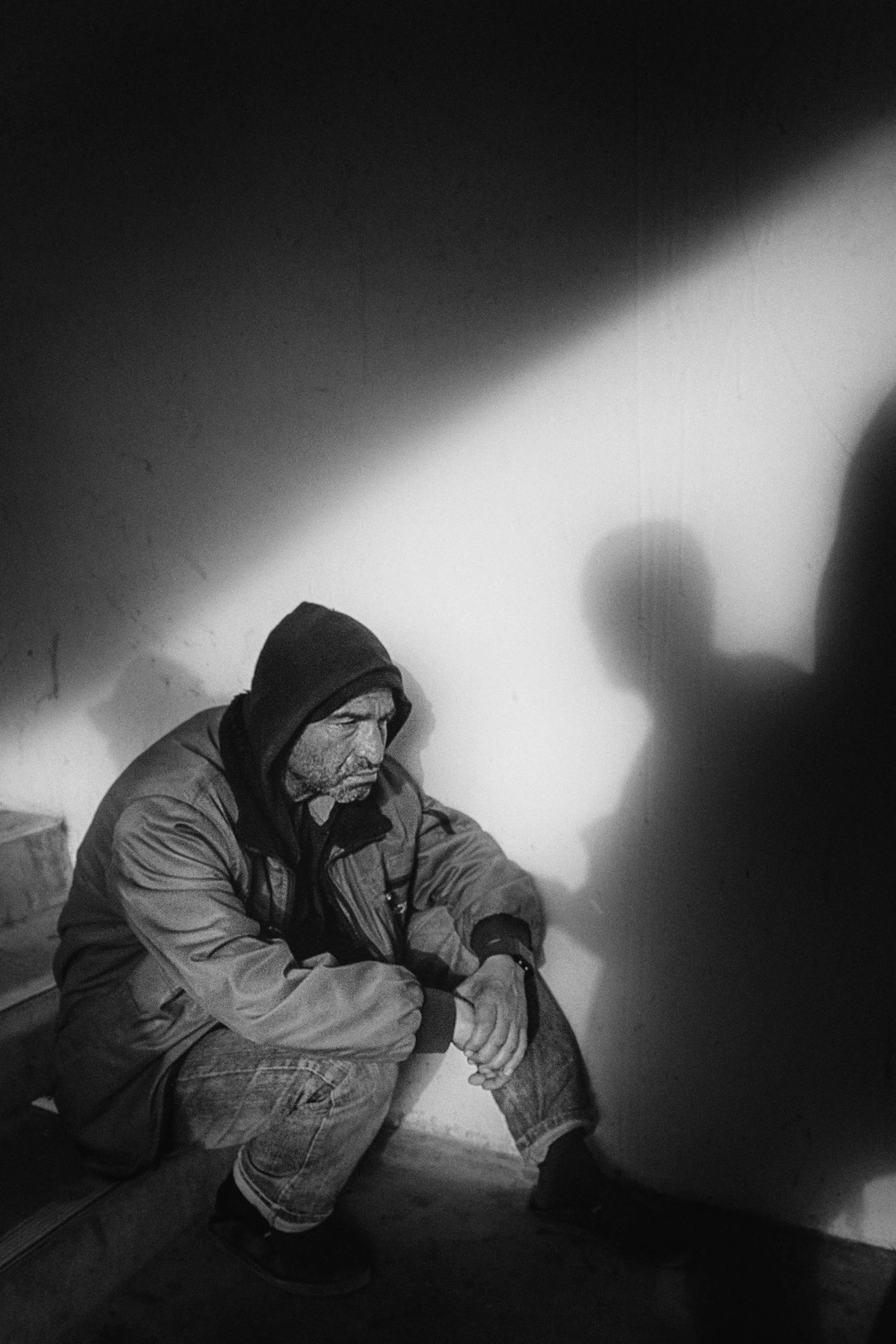 A man with a hoodie and jacket, sitting on a step, looking down with his hands clasped and resting on his knee, in a dimly lit room with shadows on the wall.