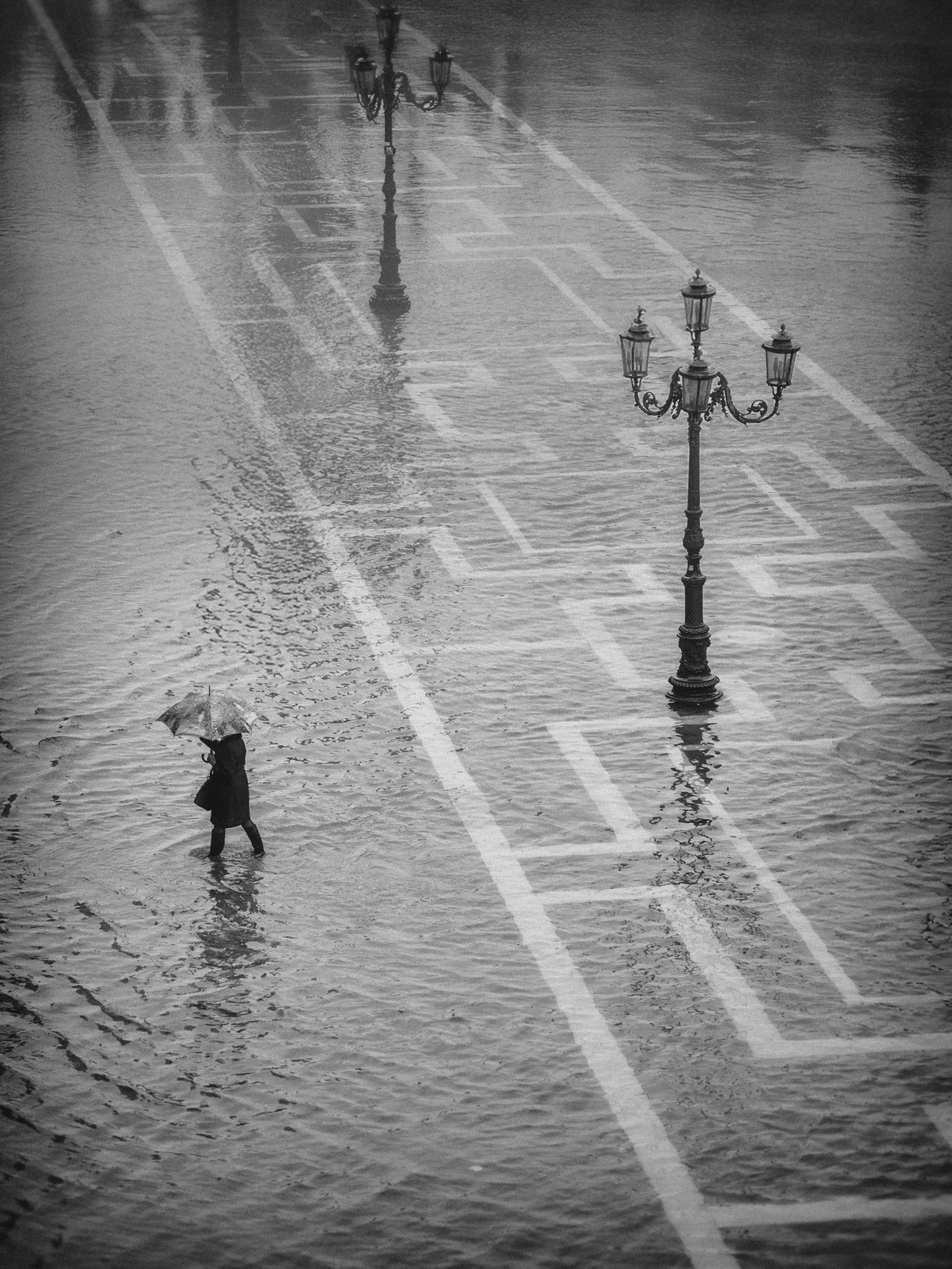 A person walking with an umbrella through a flooded street with street lamps and visible tire tracks.