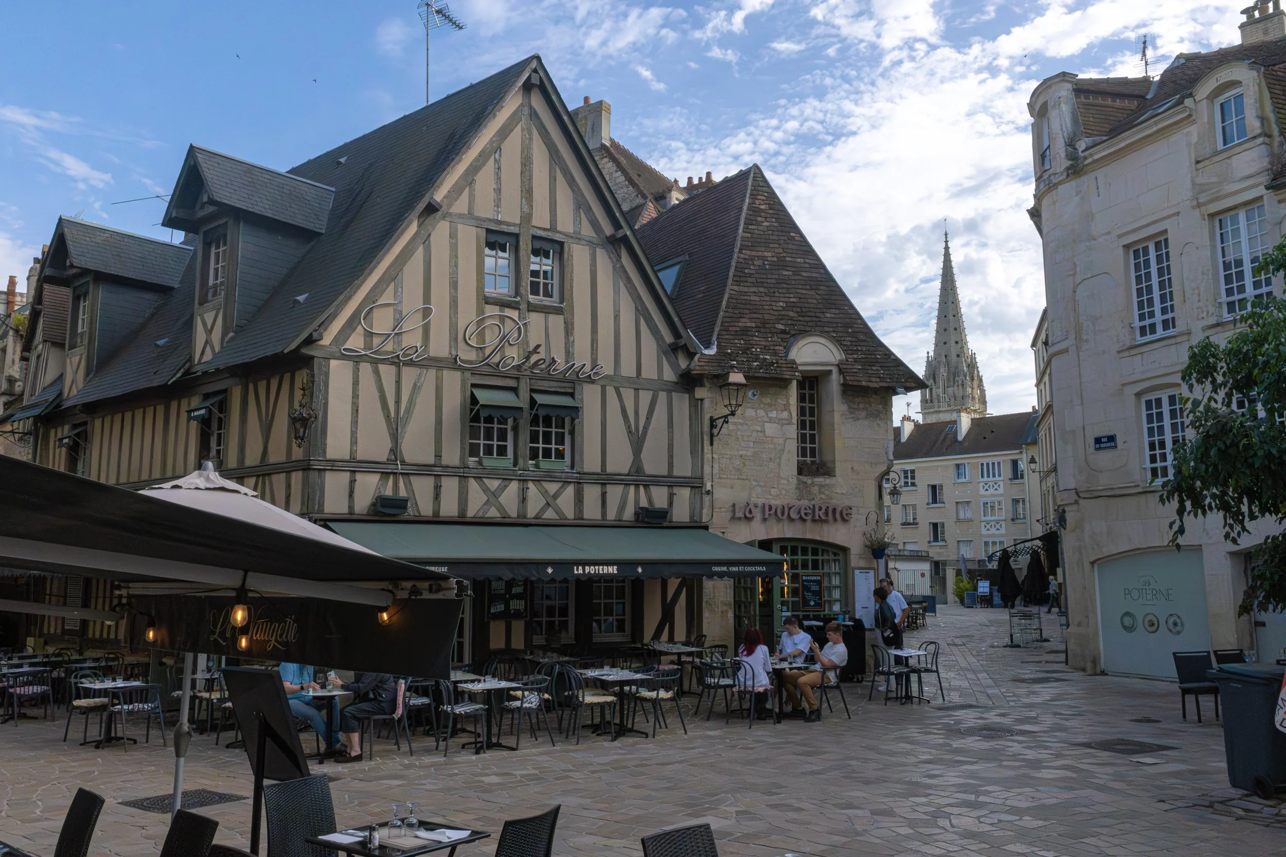 Outdoor café with tables and chairs on a cobblestone square, surrounded by historic buildings and a church steeple in the background, under a partly cloudy sky.