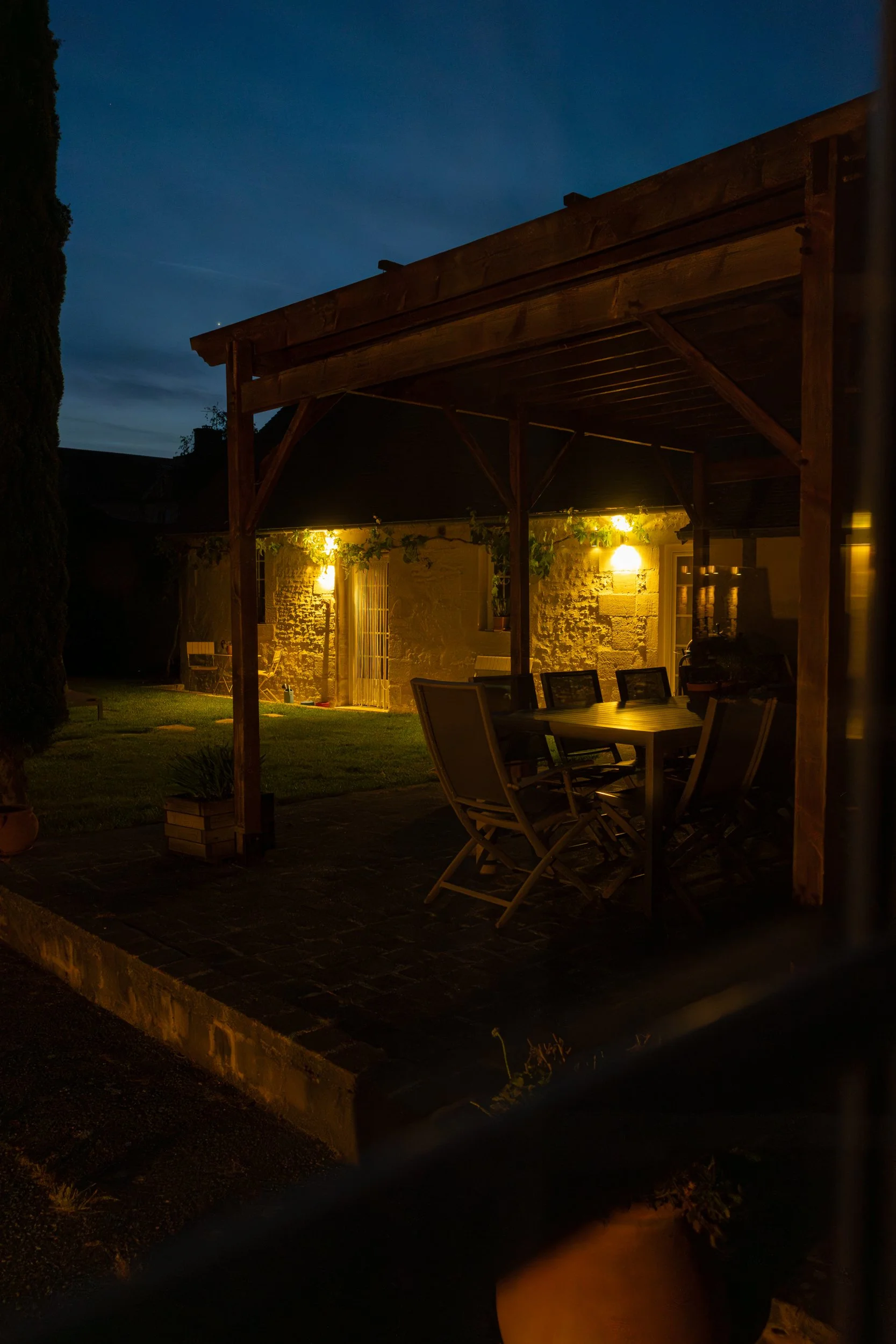 Nighttime scene of a house with a stone wall and a wooden patio cover, illuminated by outdoor lighting, with a table and chairs in the foreground and a dark sky overhead.