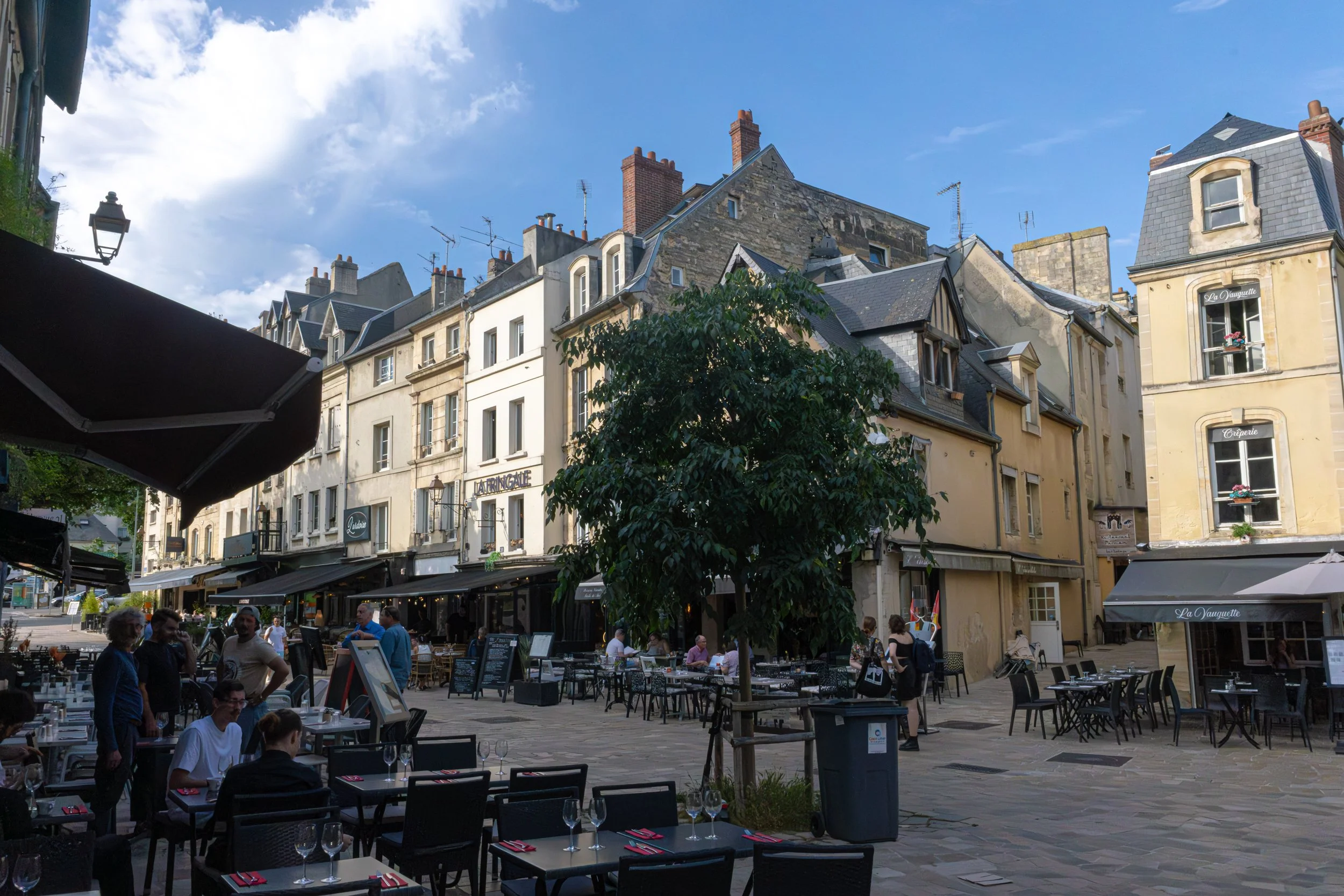 Outdoor cafe with tables and chairs, people dining, buildings with European architecture, and a blue sky with some clouds.
