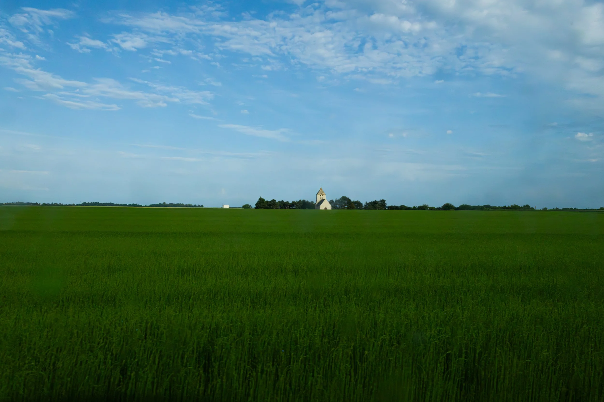 A wide green field with a small white church in the distance under a cloudy blue sky.
