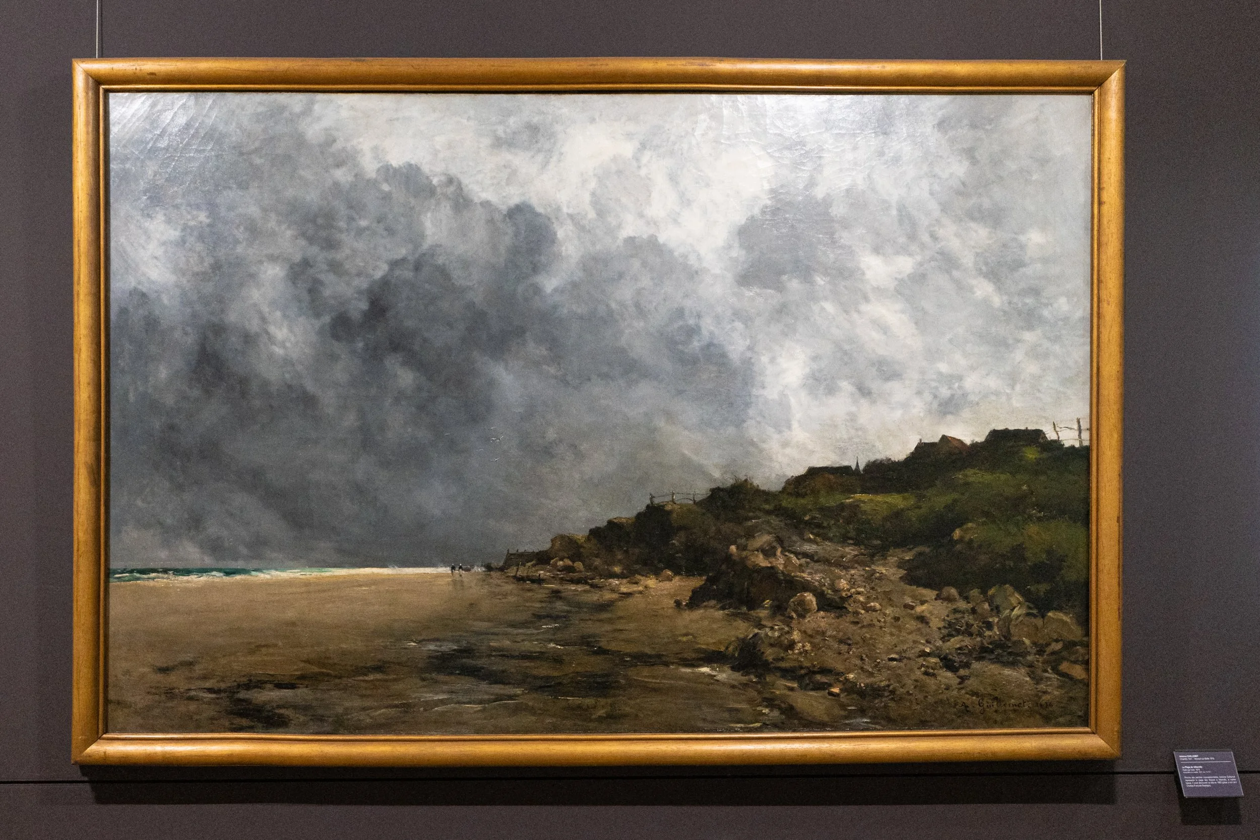 Painting of a cloudy sky over a rocky coastline with a few small figures walking on the beach.