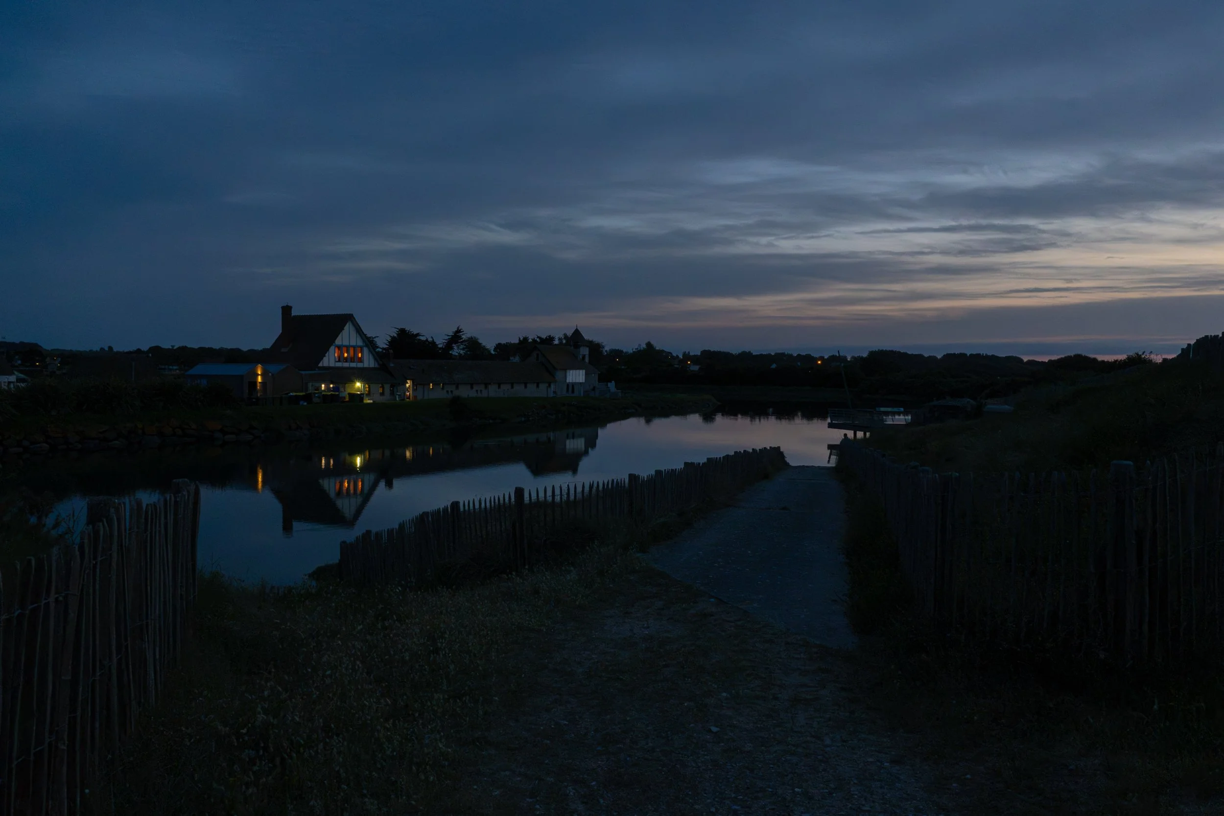 A calm body of water with reflections of houses and trees, dark cloudy sky, and a faint sunset in the distance.