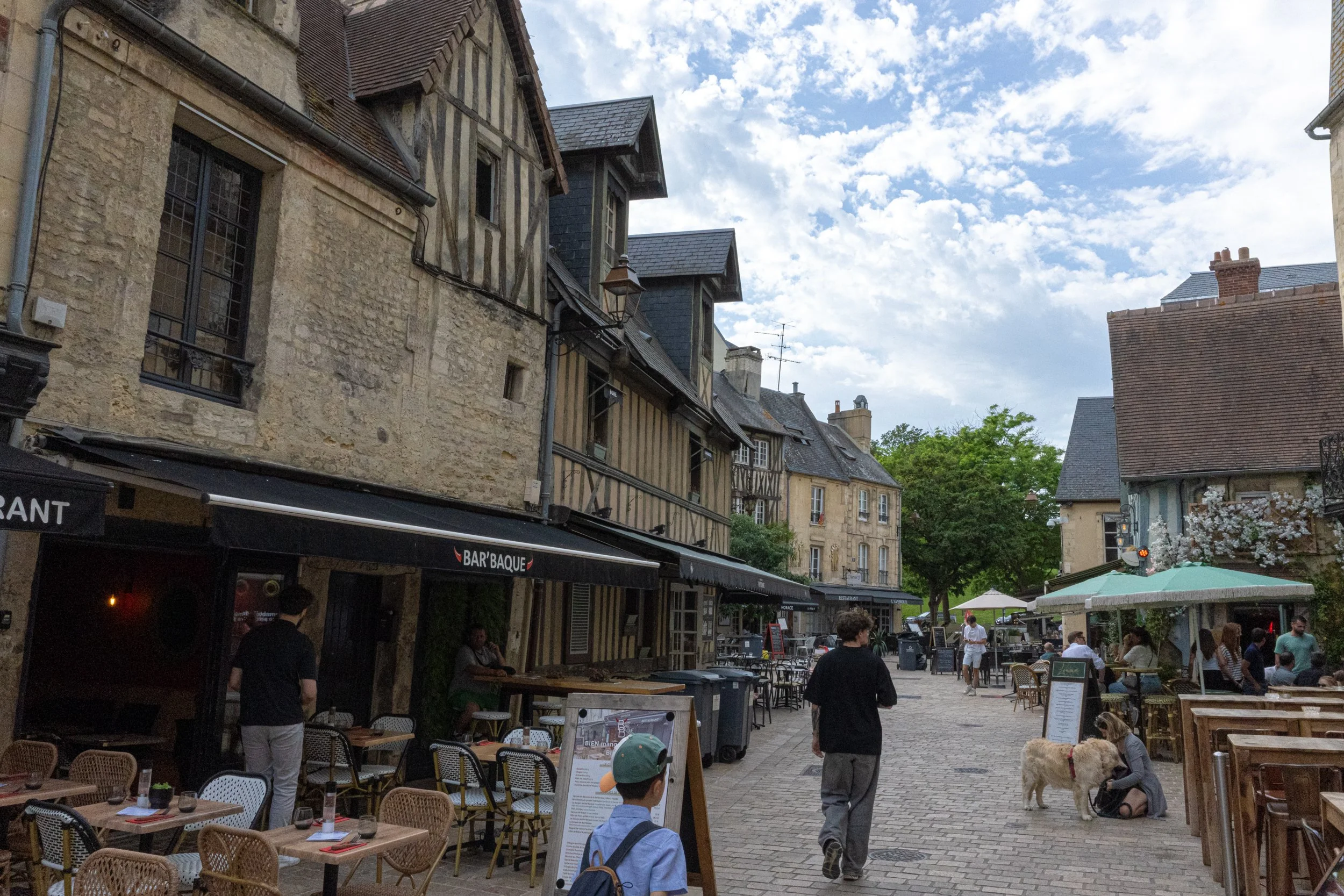 Outdoor street scene with old European-style buildings, outdoor seating, and people walking and sitting. Some trees and a cloudy sky are visible.