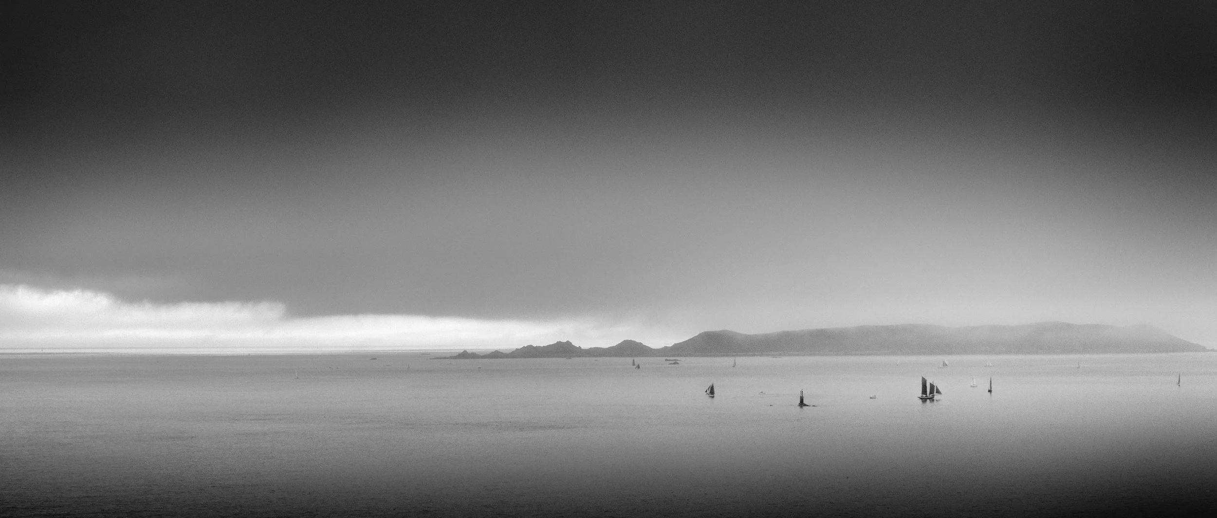 Black and white photo of a seascape with several small sailboats on the water and a distant island or landmass in the background under cloudy skies.