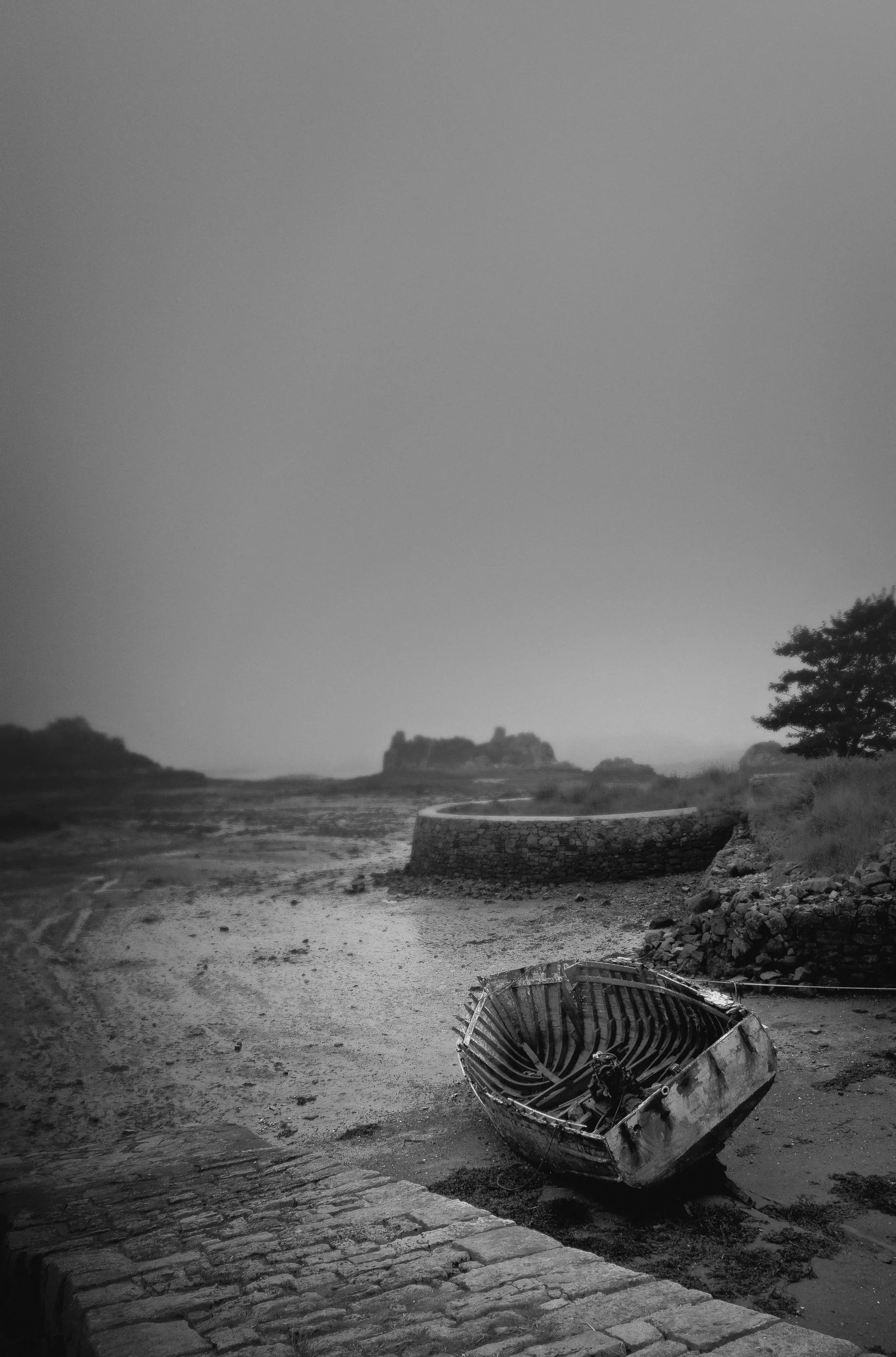 A black and white photo of a fallen boat on a muddy riverbank with an old stone pathway leading towards the water, with trees and a distant island in the background.