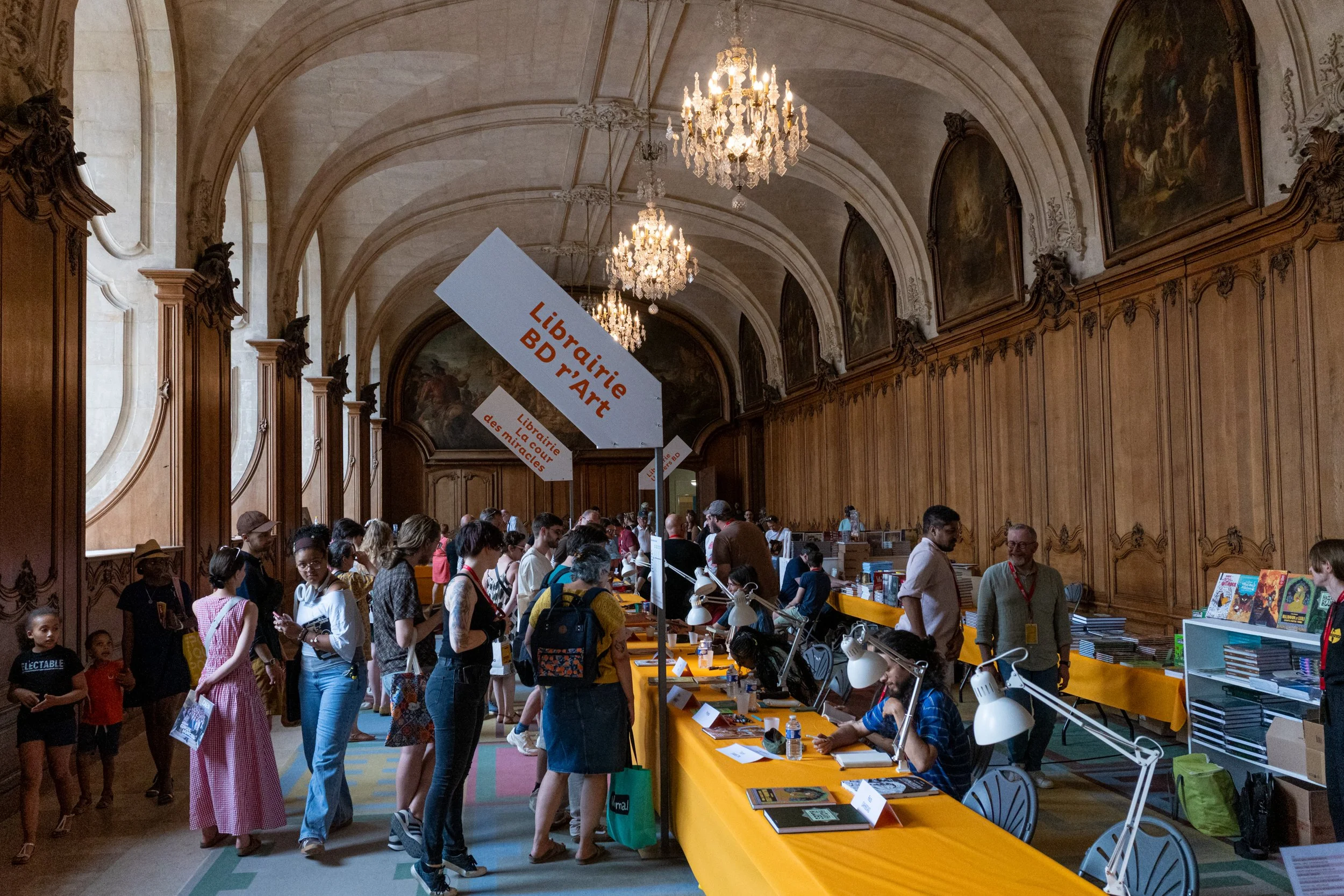 People shopping at a book fair inside a grand hall with high arched ceilings and chandeliers. There are books and signs on tables, with many customers browsing and interacting with vendors.