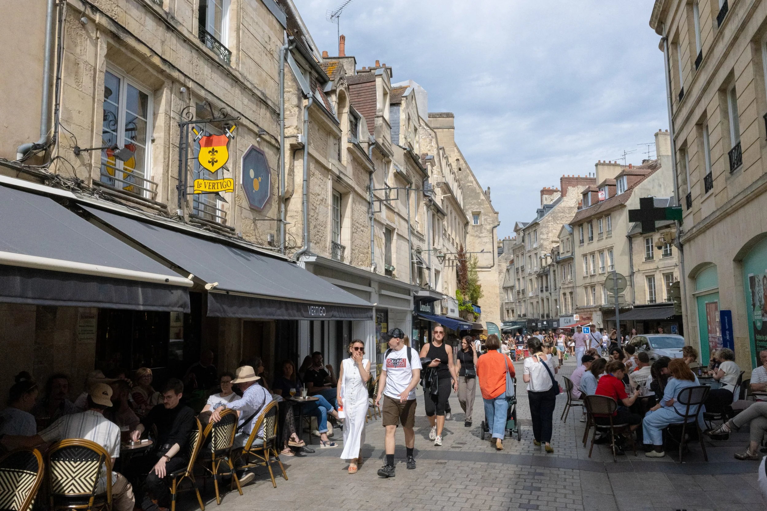 A bustling street scene with outdoor café seating and people walking, with historic stone buildings and various signs, including a green cross indicating a pharmacy, under a partly cloudy sky.
