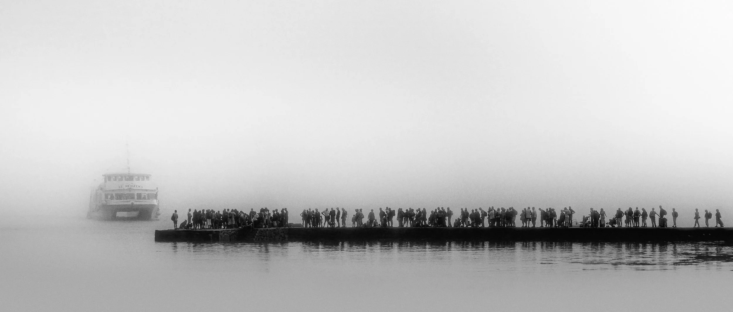 A foggy scene at a dock with a large group of people standing on the pier, and a boat in the background partially obscured by the fog.