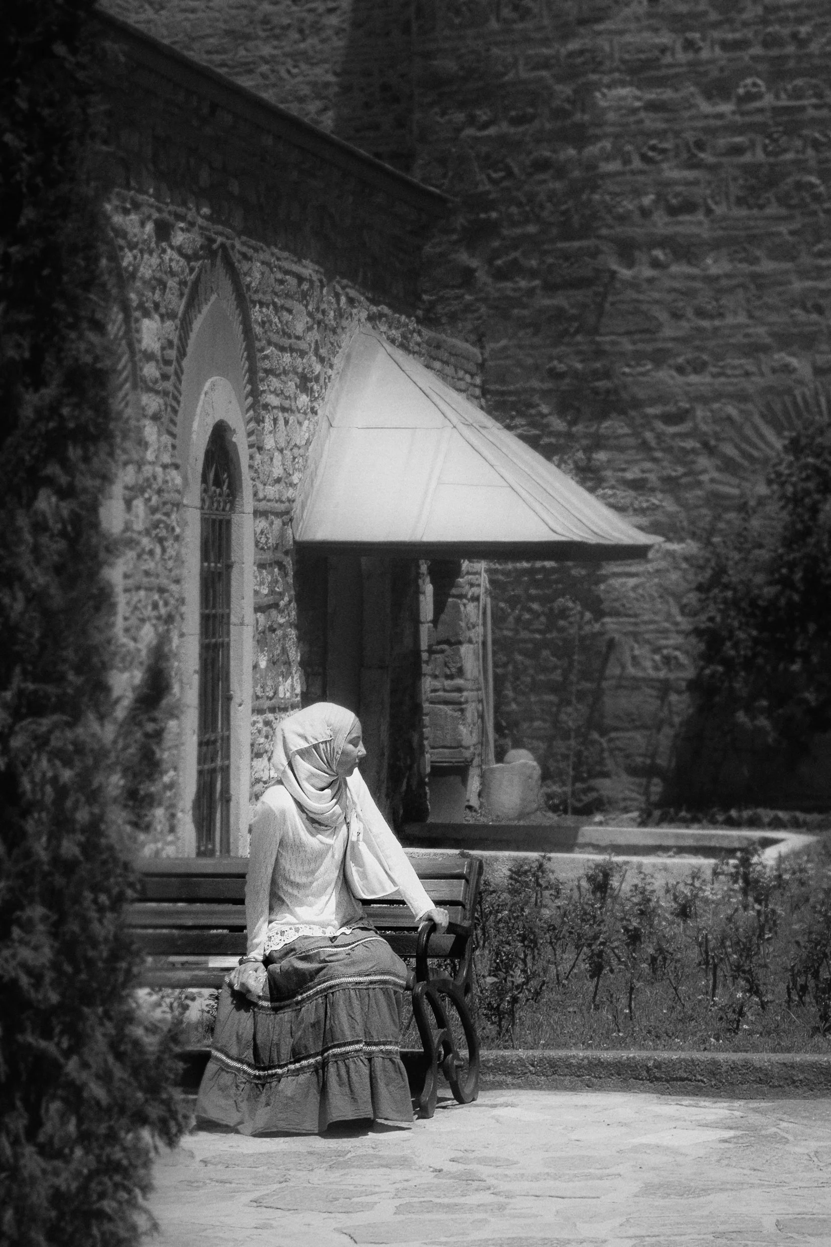A woman sitting on a bench outside a stone building, wearing a headscarf and a long dress, with an umbrella and plants nearby.