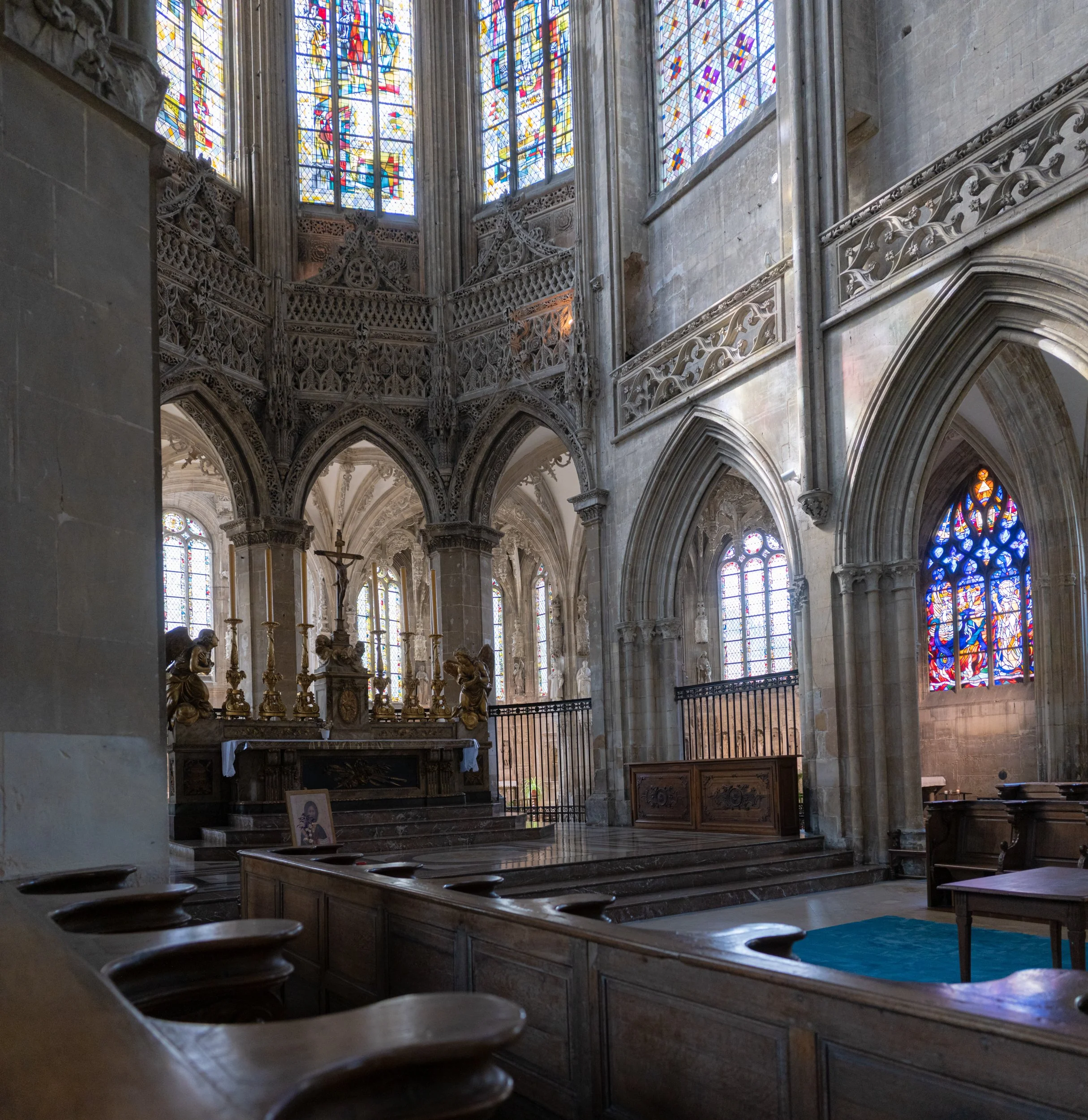 Interior of a Gothic-style church or cathedral with stained glass windows, stone arches, ornate carvings, and a wooden altar with religious statues and candles.