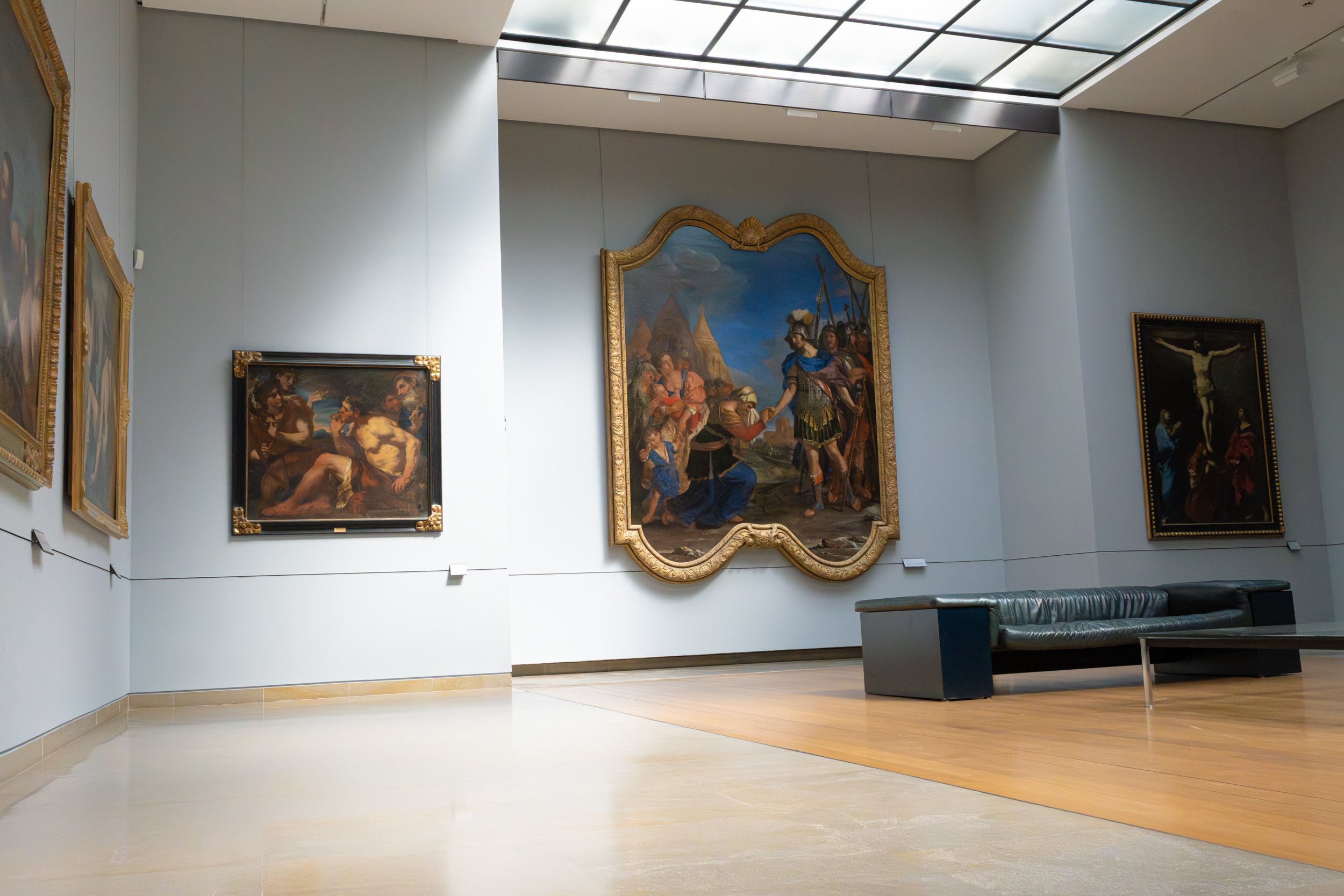 Art gallery with ornate framed historical paintings on light blue walls, modern ceiling with skylights, wooden flooring, and a black sofa in the foreground.