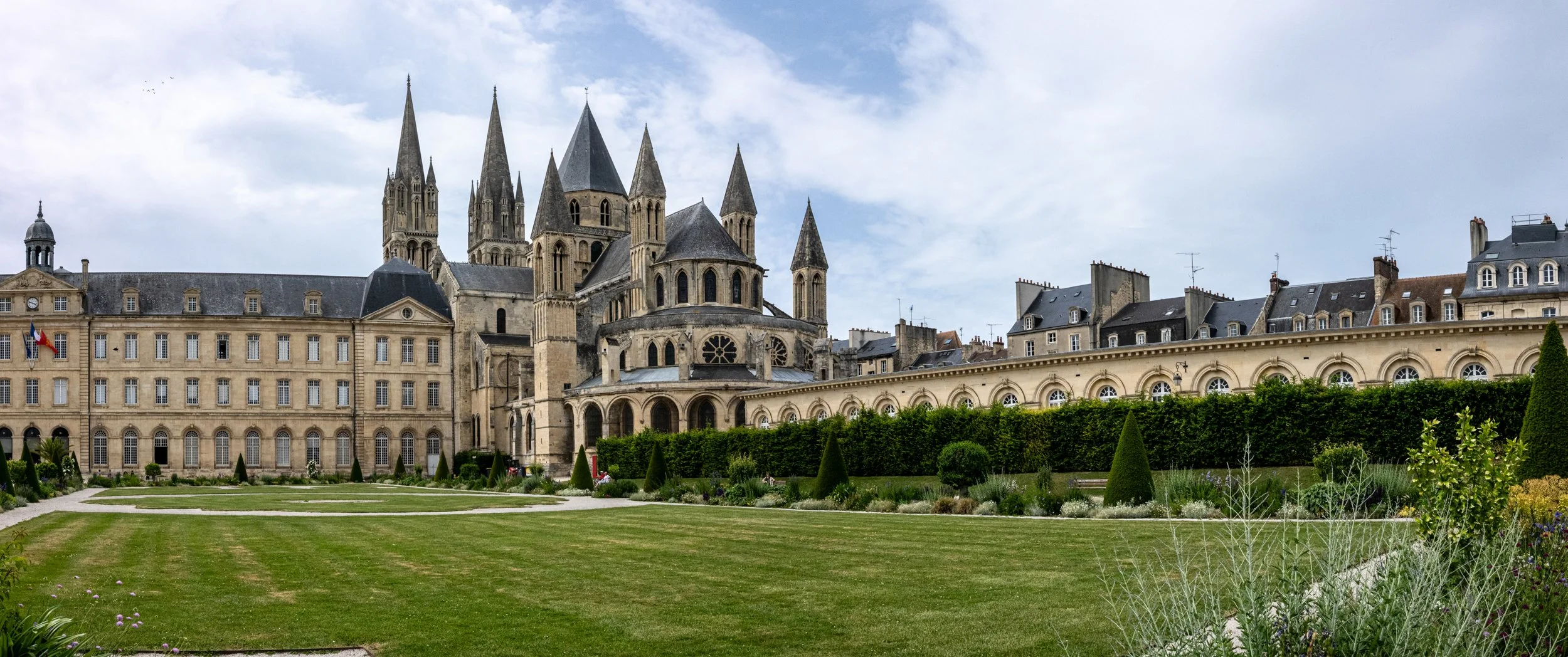 Historical European-style cathedral with multiple tall spires, surrounded by a well-maintained garden and classic stone buildings, under a partly cloudy sky.