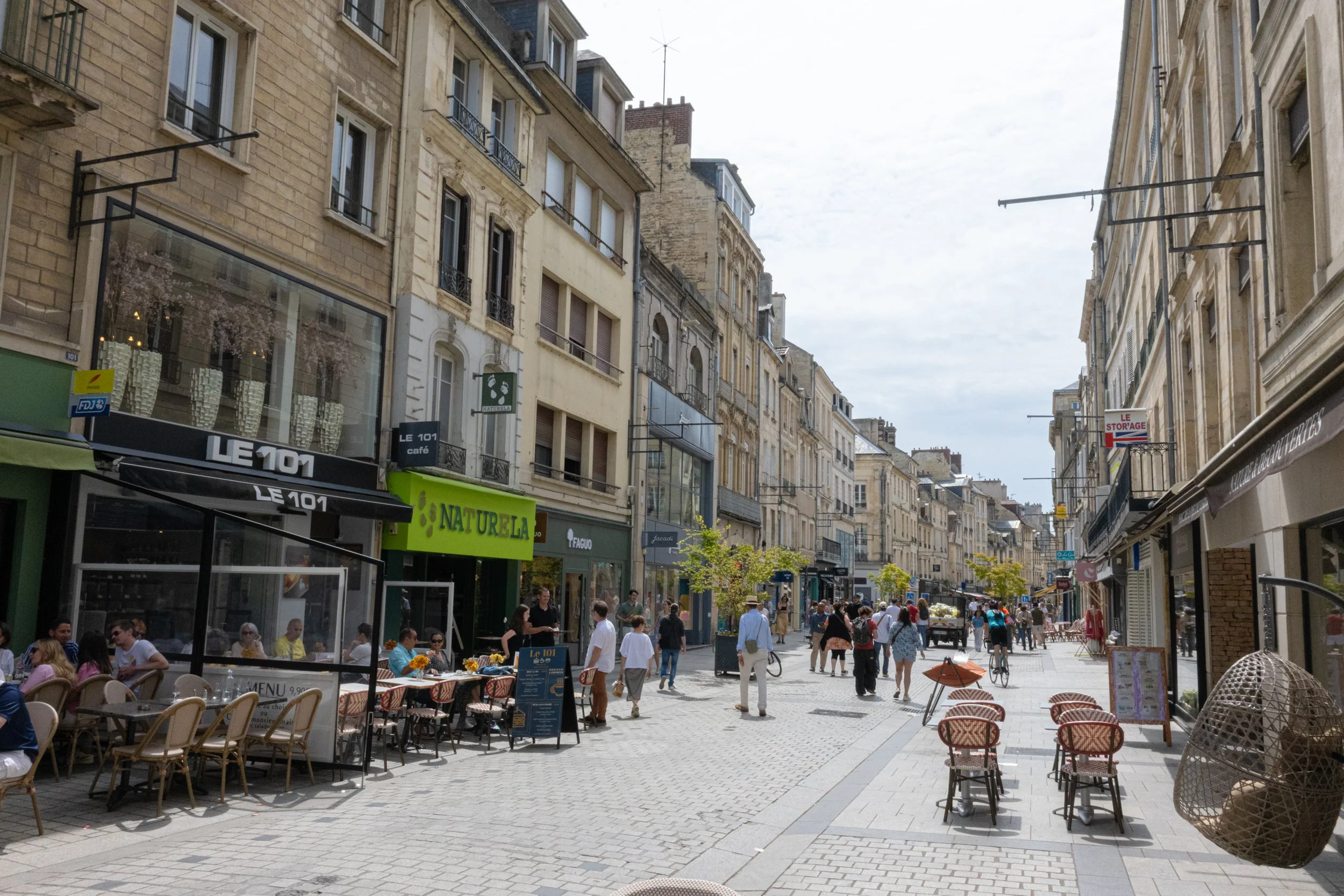 A busy outdoor street scene with cafes and shops, people walking and sitting at tables, and historic buildings lining the street on a partly cloudy day.