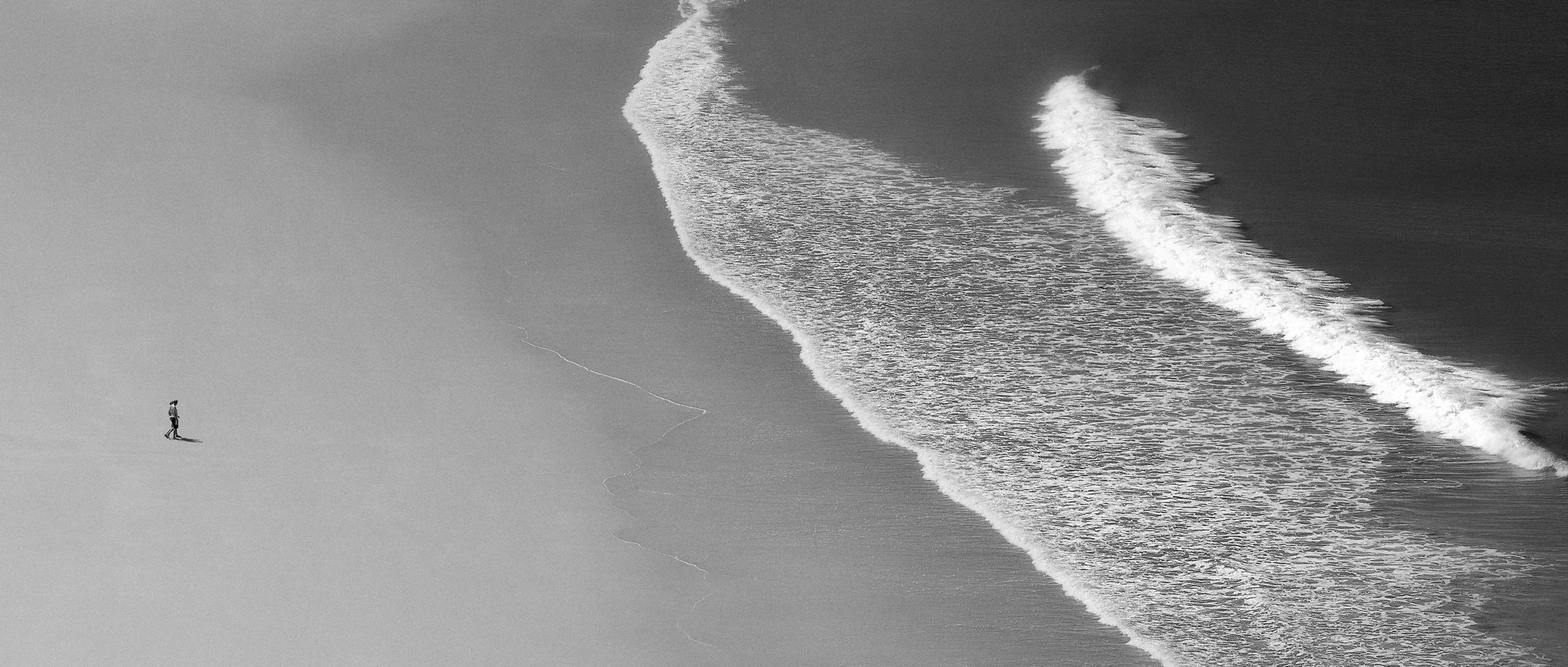 Black and white photo of a person walking along a sandy beach, with ocean waves rolling onto the shore in the background.