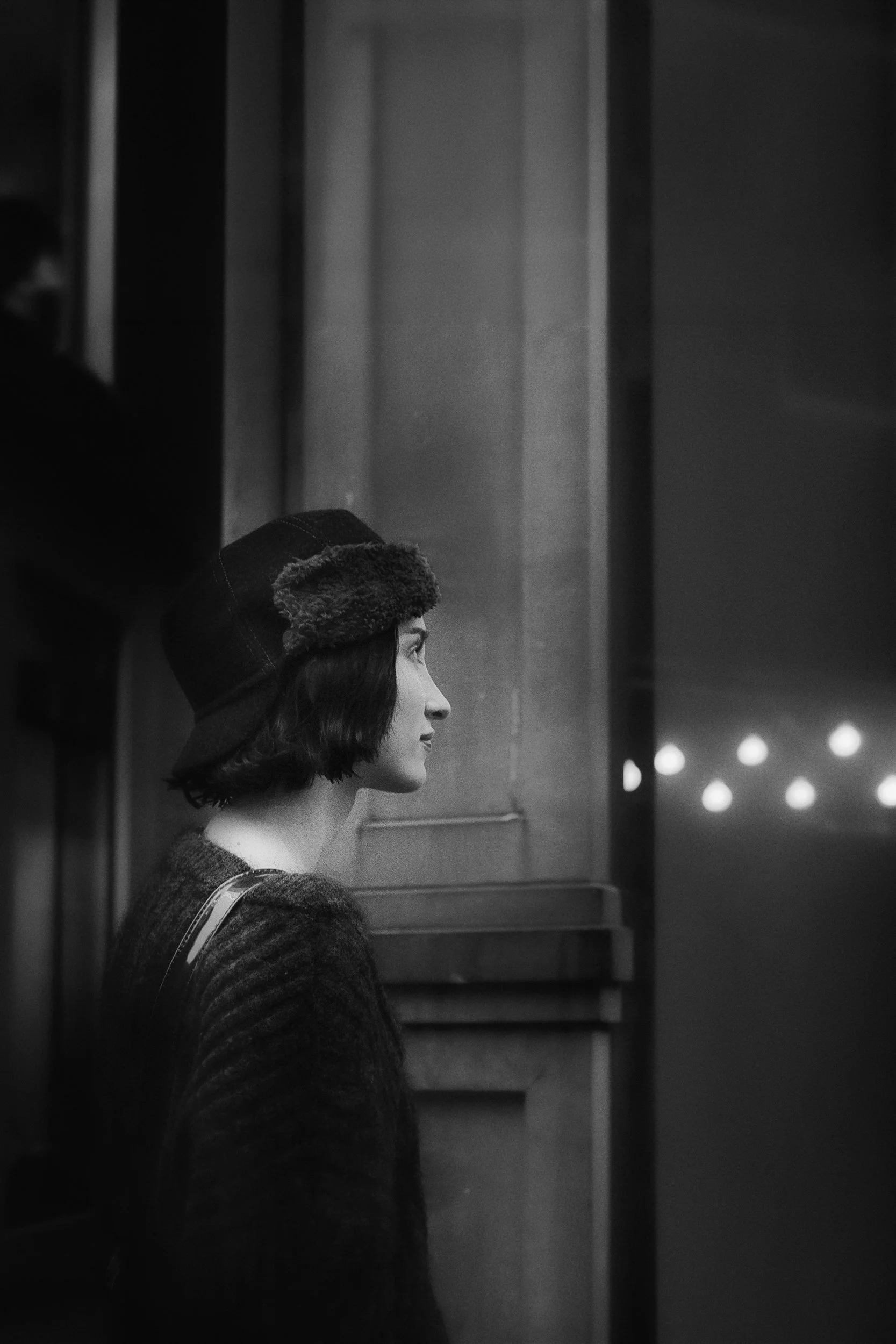 Black and white photo of a woman with dark, wavy hair, wearing a coat and a fur hat, standing in front of elevator doors.