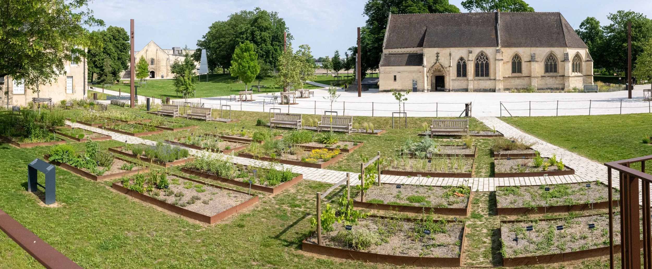 A historic church with Gothic windows and a stone facade, surrounded by a community garden with raised beds and pathways, trees, and benches.