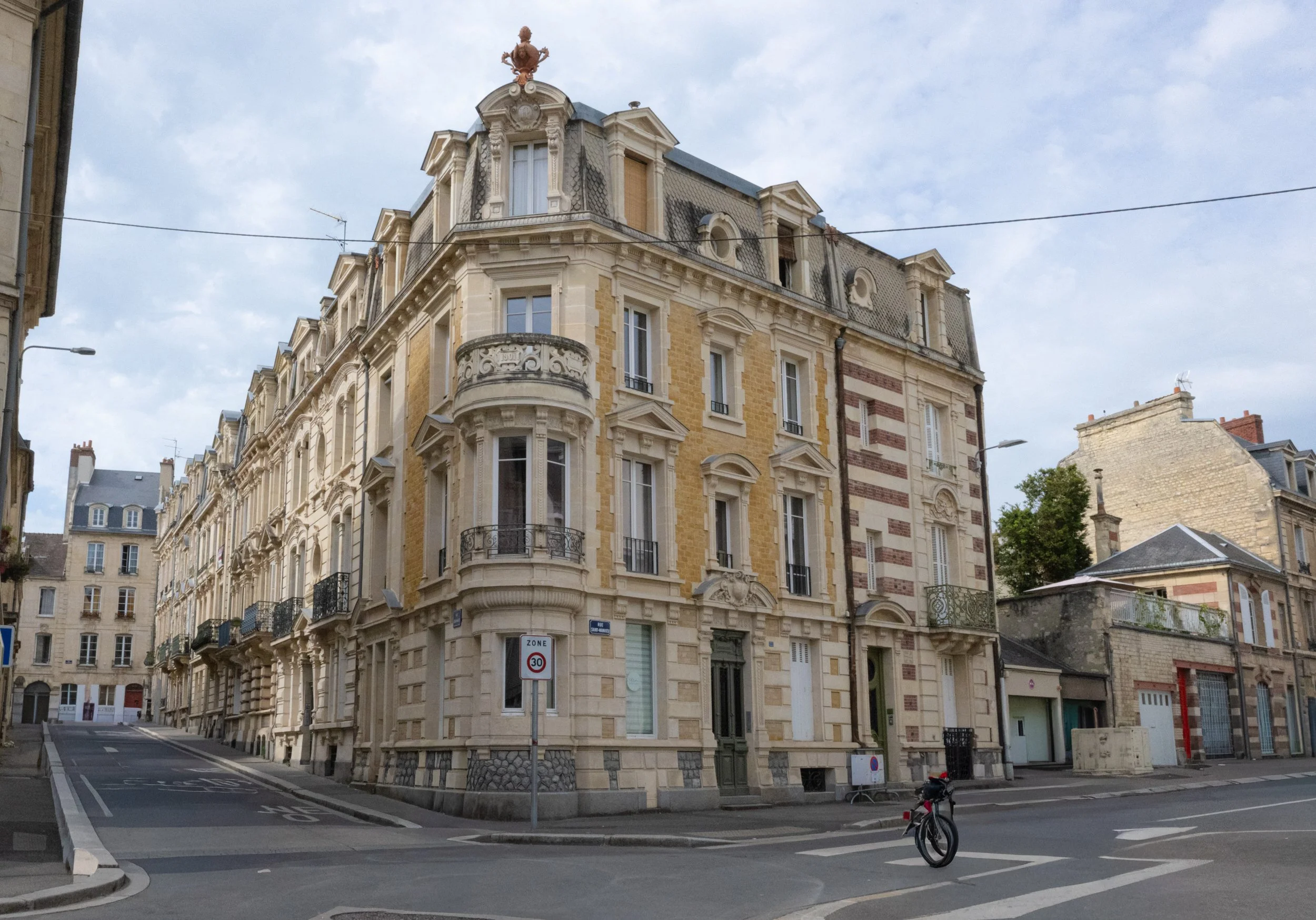 A corner building in a European city with ornate architecture, including decorative moldings, balconies, and a statue on the roof, with narrow streets and parked bicycles in the foreground.