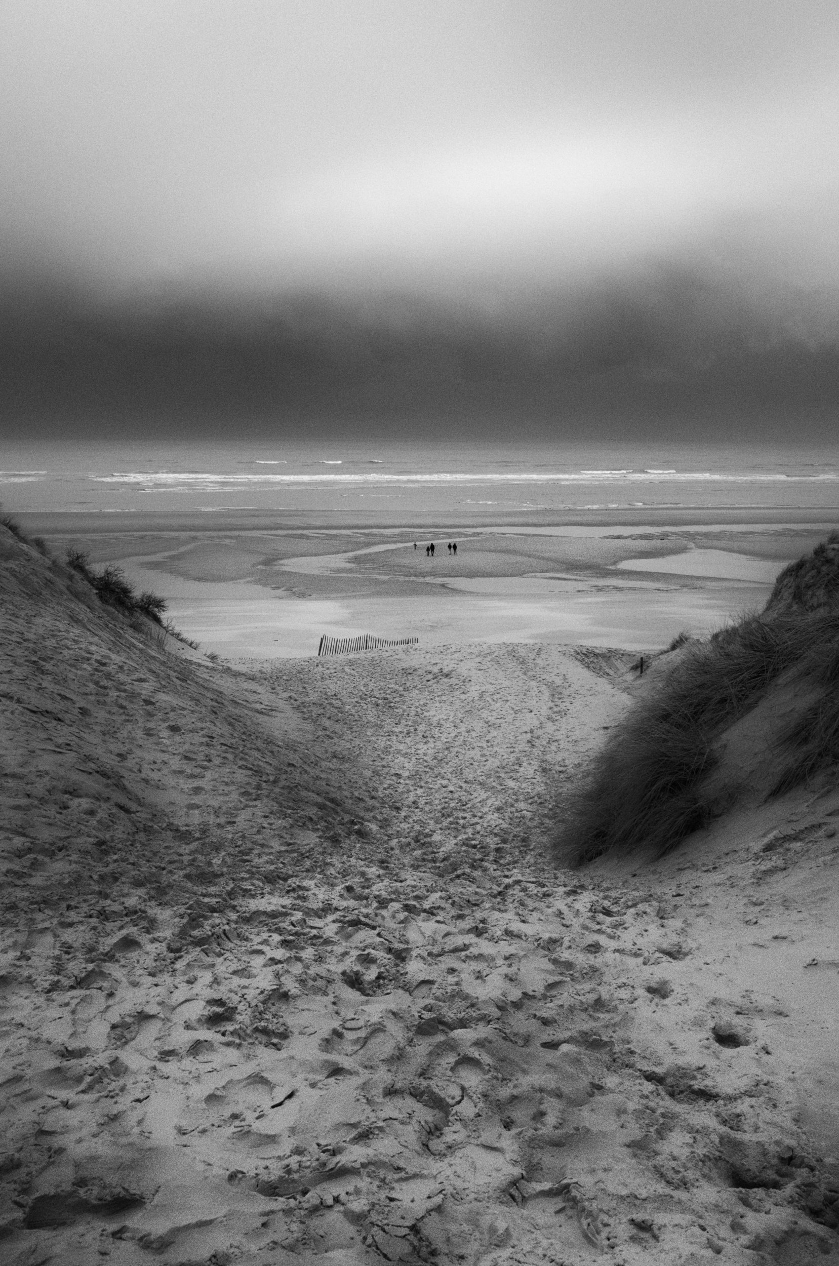 Black and white photograph of a beach with sand dunes in the foreground, a small group of people walking near the water, and a cloudy sky above.