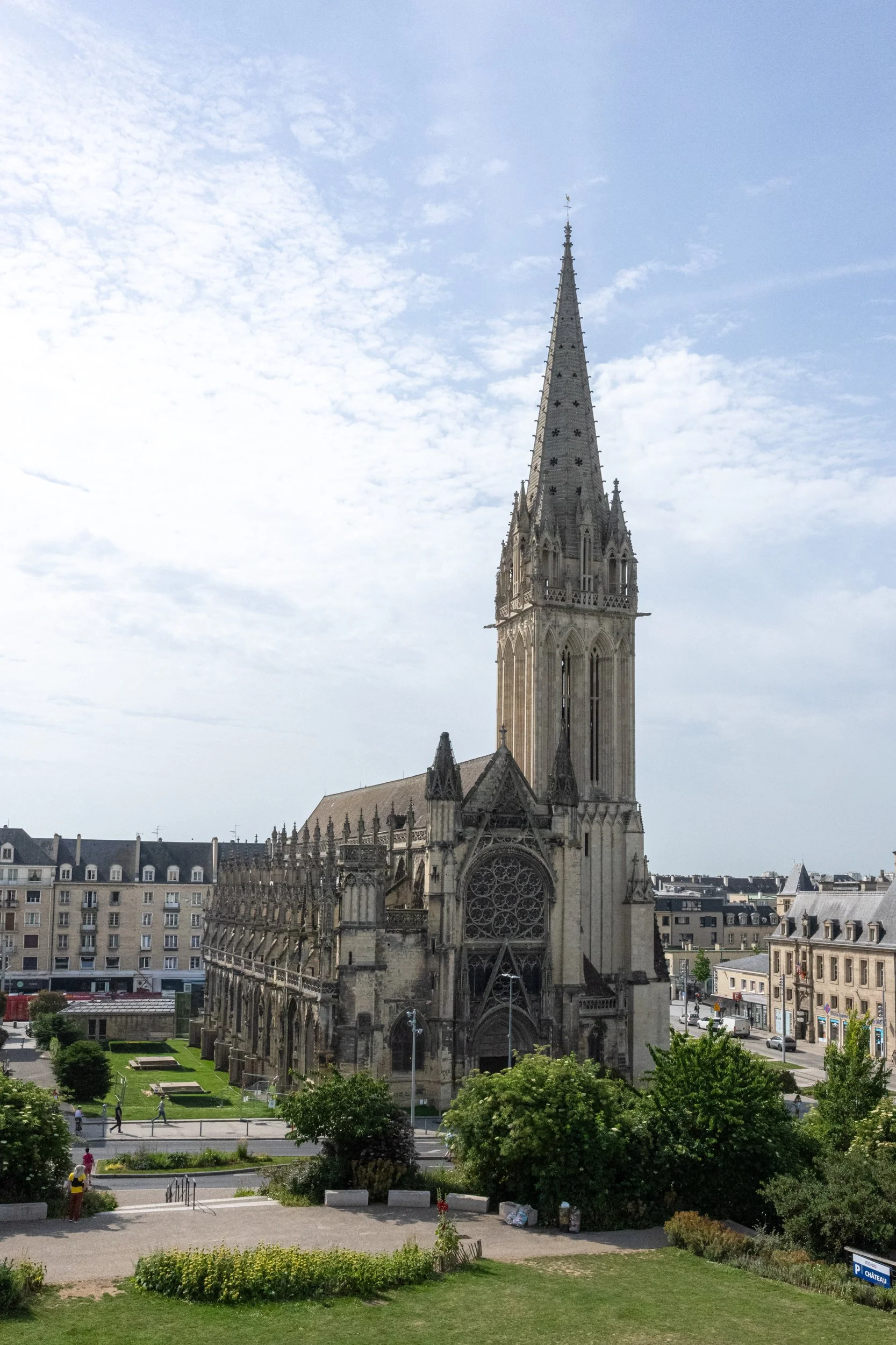 A large Gothic cathedral with a tall spire, intricate stonework, and rose windows. The church is surrounded by trees, houses, and a town with people walking nearby.