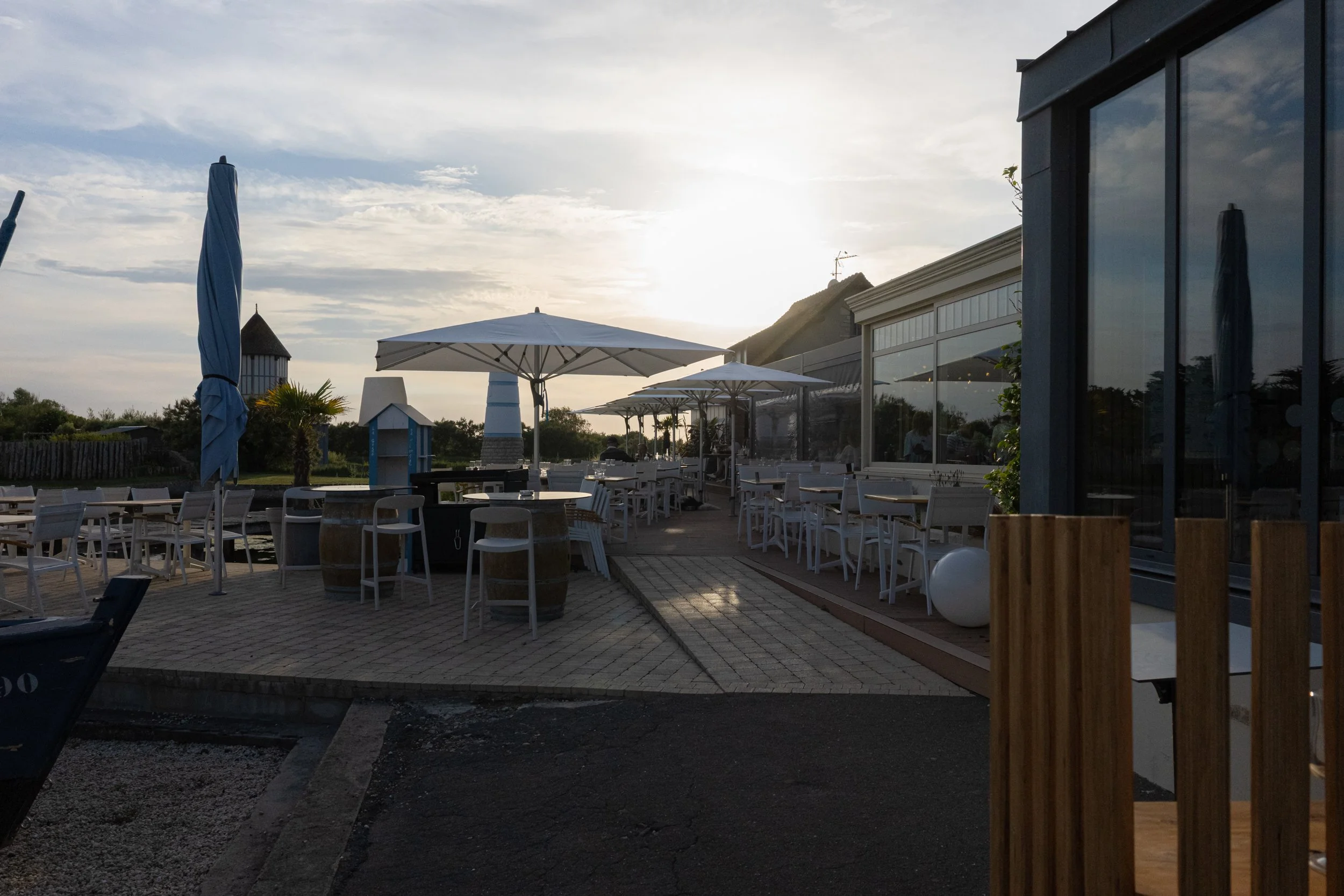 An outdoor dining area with white tables, chairs, umbrellas, and a modern building with large glass windows. The sun is setting, creating a bright glow in the background. There are some trees and a small structure with a thatched roof in the distance