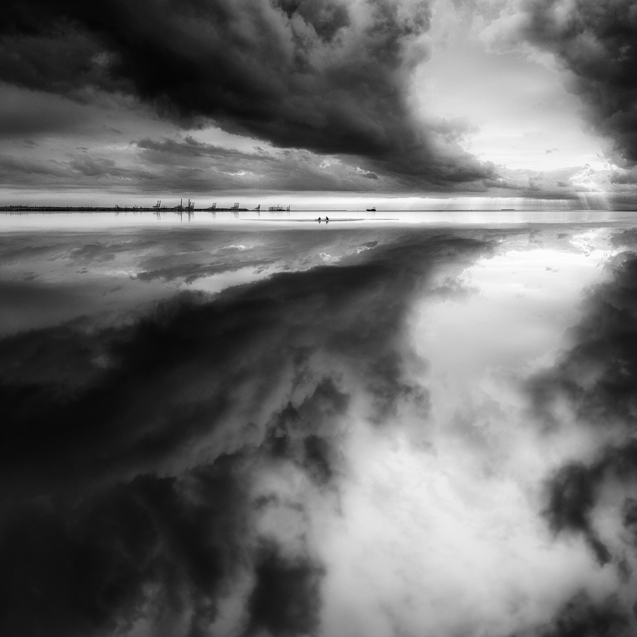 Dark storm clouds over a calm body of water with boats and industrial cranes in the distance.