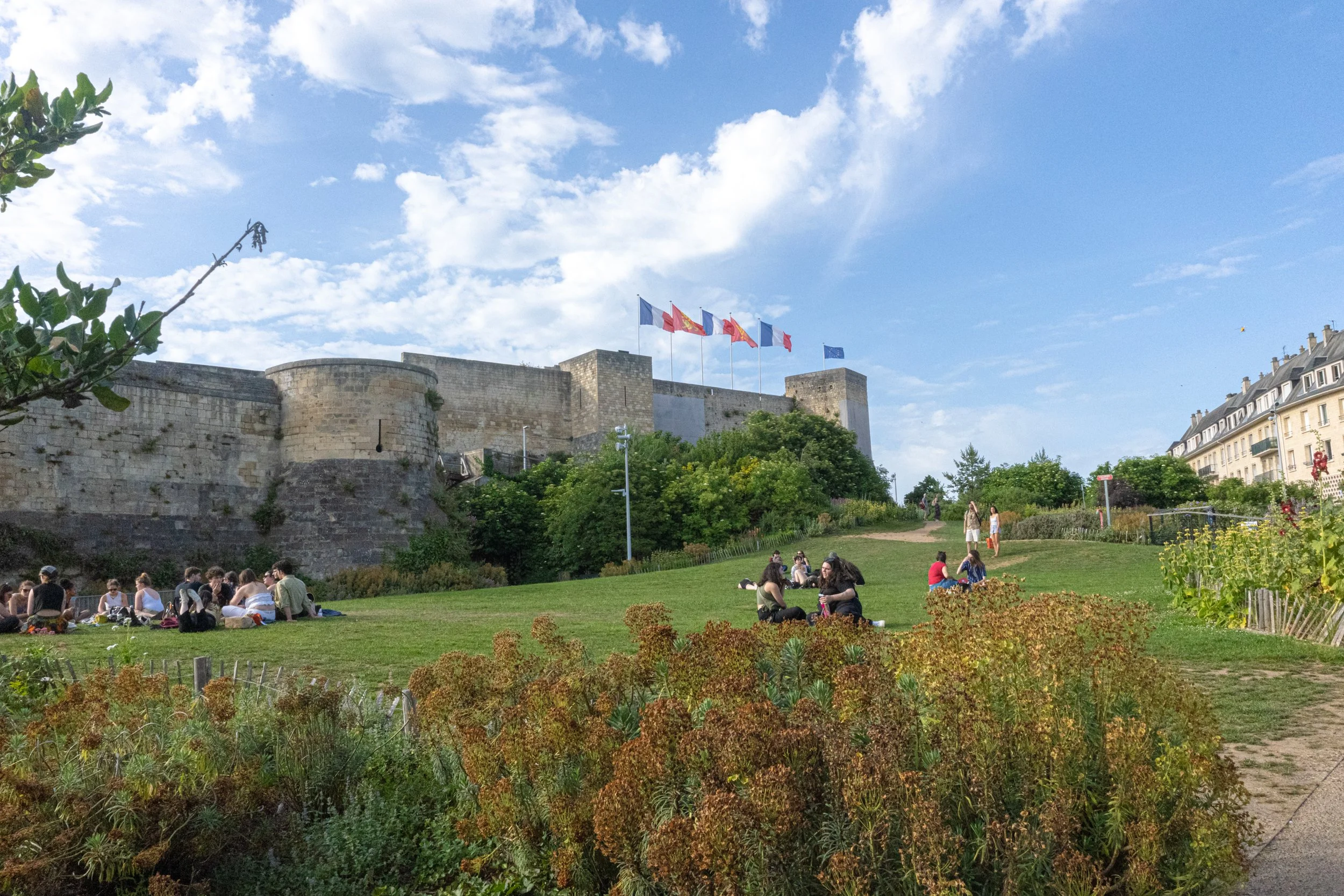 People sitting on a grassy park with flowers, trees, and a historic stone fortress in the background flying French flags under a blue sky with clouds.
