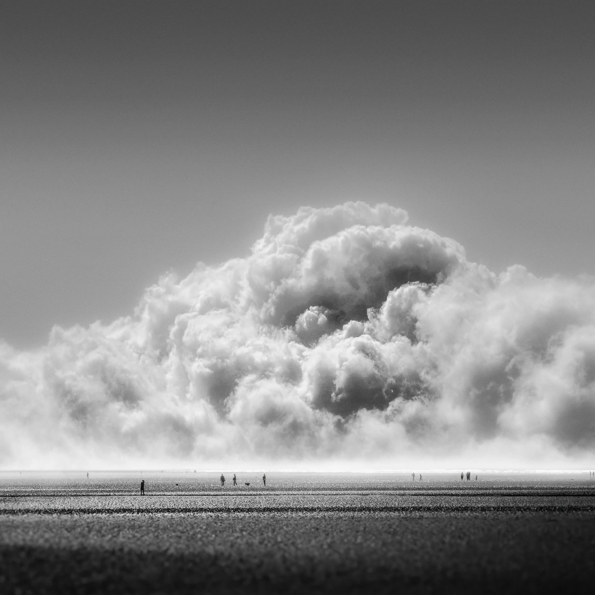 Black and white photograph of a beach with small silhouetted figures walking along the shore and clouds overhead.