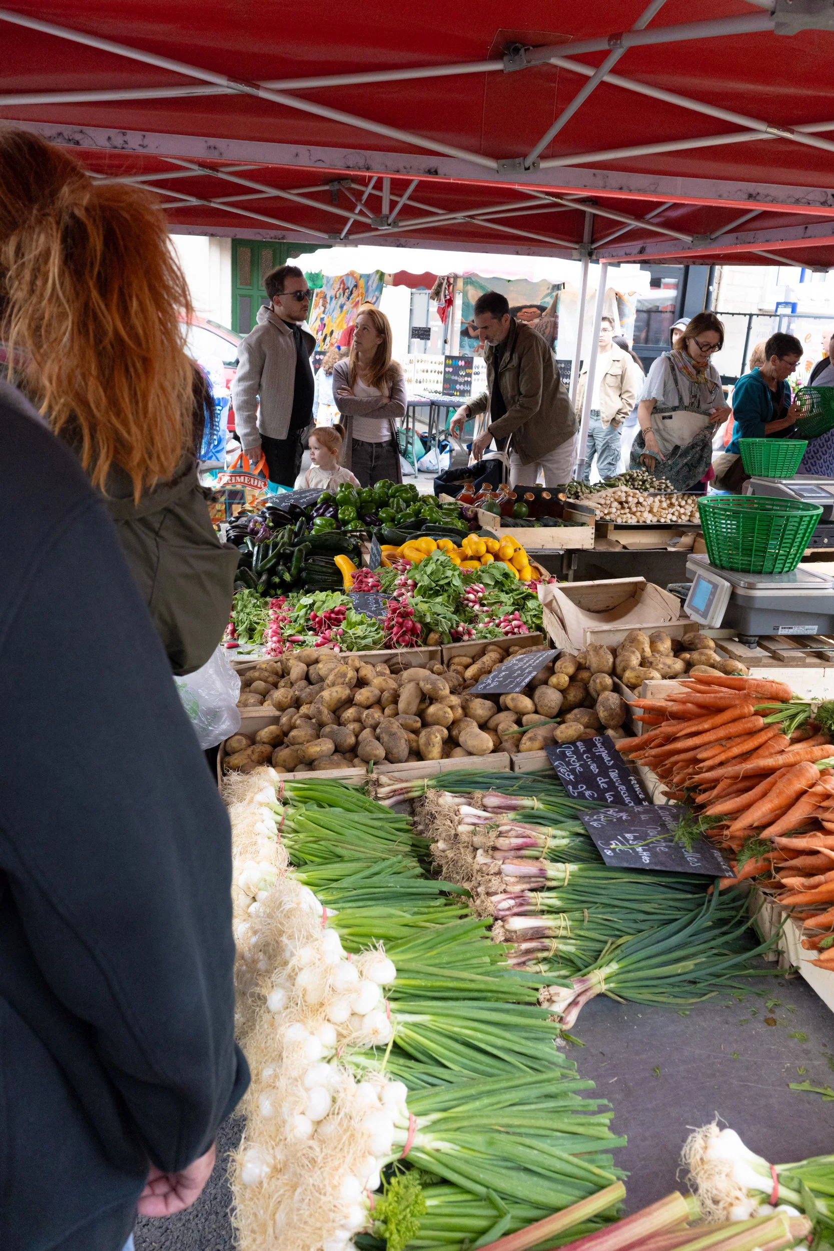 View of a farmers market stall selling fresh vegetables including potatoes, carrots, green onions, radishes, garlic, and peppers, with people browsing and shopping under a red canopy.