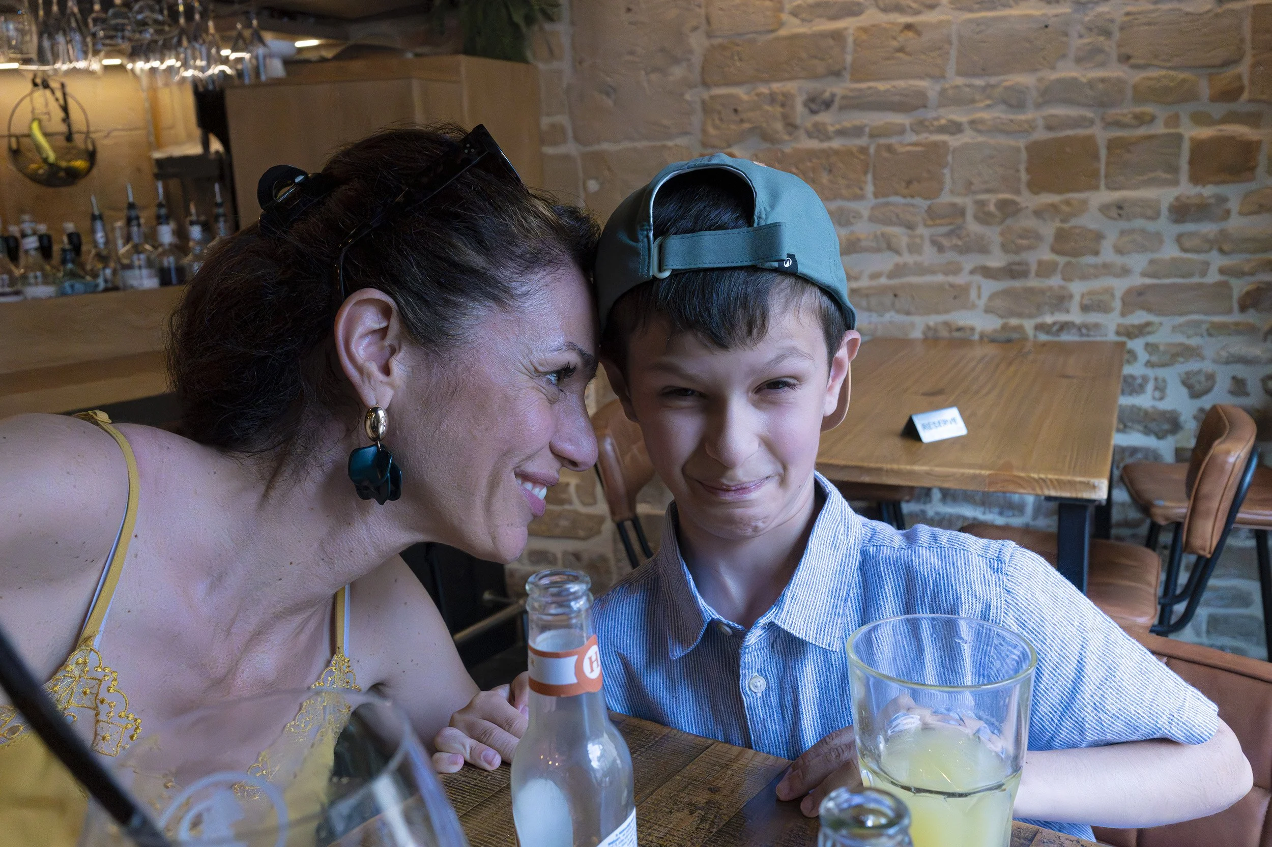 A woman and a young boy sitting at a wooden table, taking a selfie together. The woman is smiling, wearing earrings and a yellow dress with lace details. The boy is wearing a blue cap backwards and a striped shirt, smiling slightly. There are drinks 