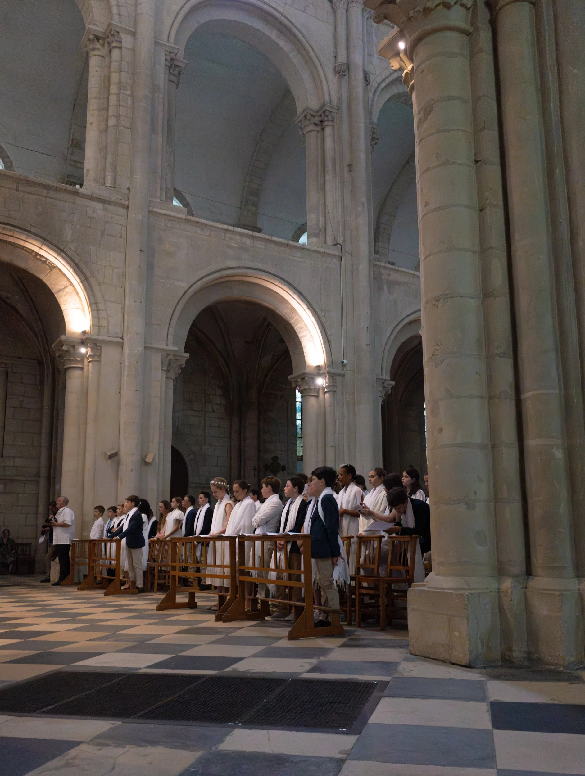 A group of people dressed in white and black robes standing in a row inside a grand stone cathedral with high vaulted ceilings, large columns, and arched windows.