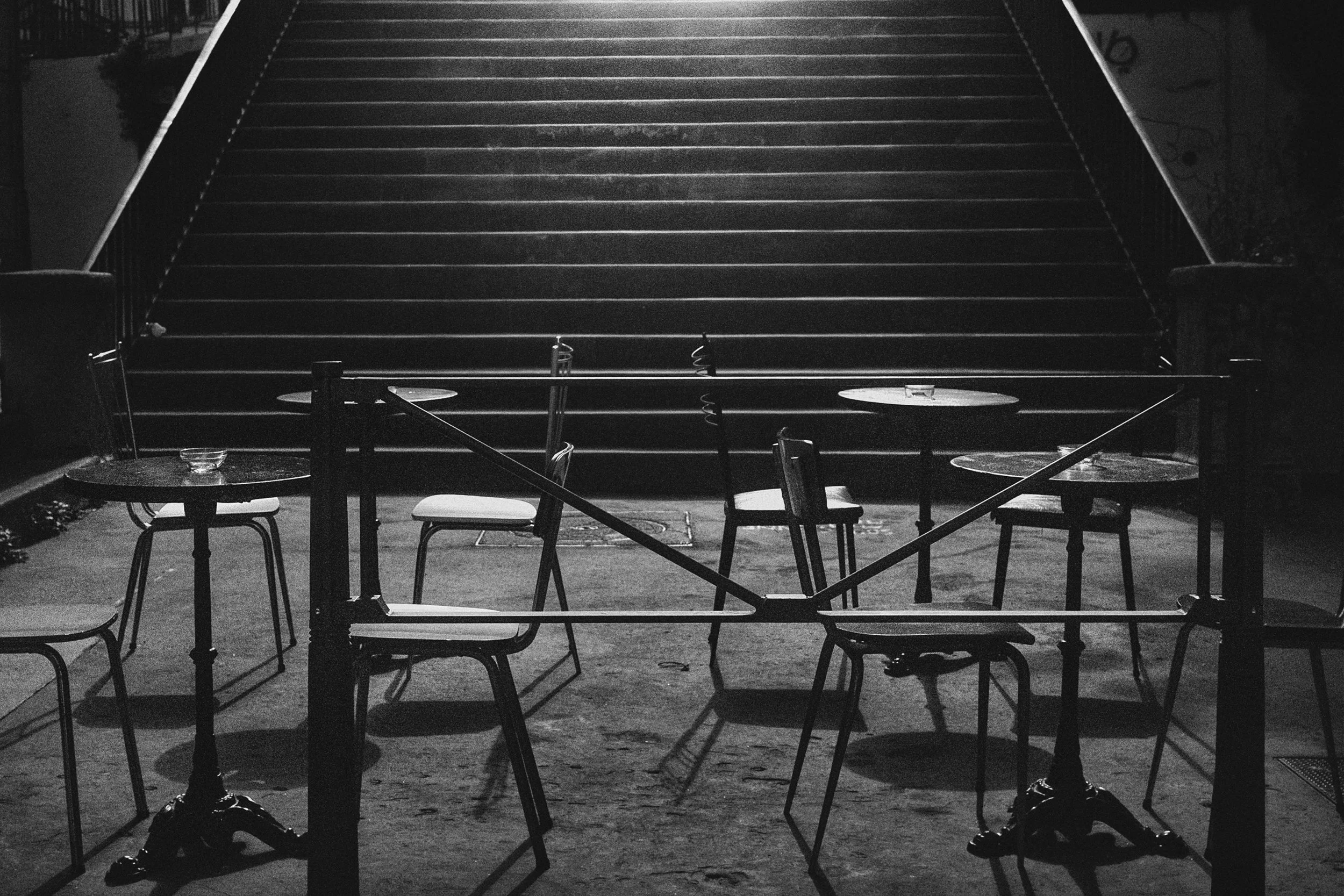 Empty outdoor seating area with tables and chairs, a staircase in the background, black and white photography.