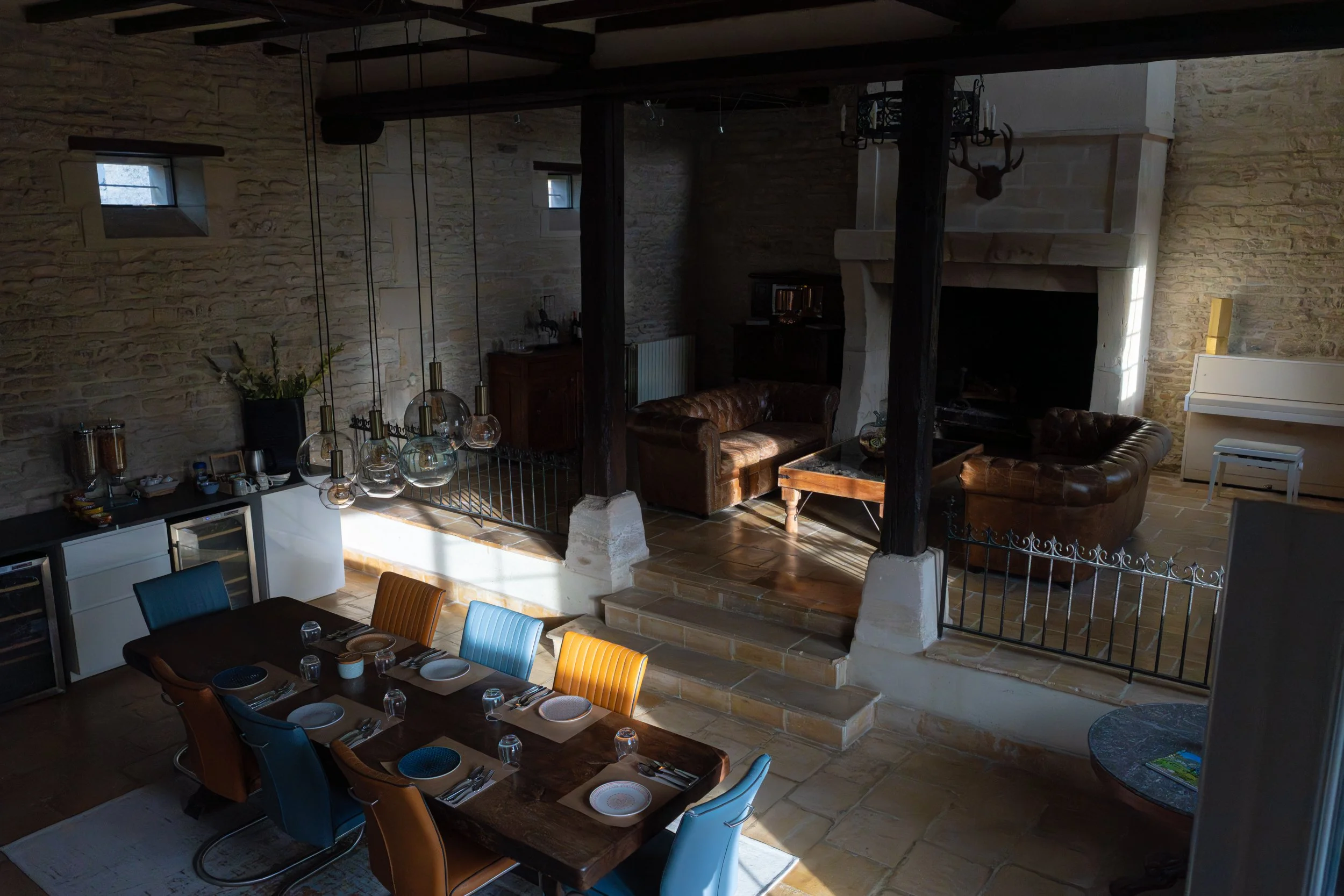 Interior of a rustic dining room with a long wooden table set with plates, glasses, and silverware, surrounded by colorful chairs, a stone wall, and a staircase with hanging pendant lights, large windows bringing natural light, and a cozy seating are