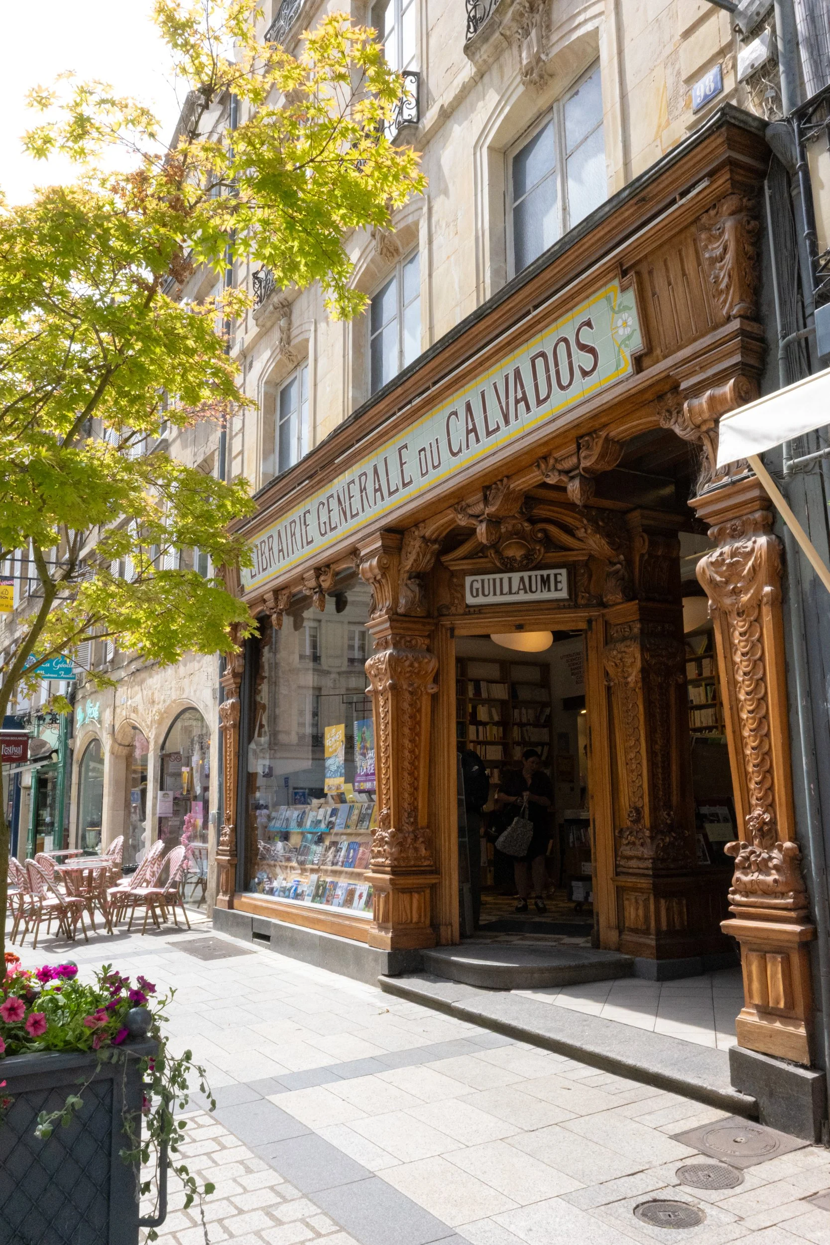 French bookstore with a wooden ornate storefront and signage that reads 'Librairie Generale du Calvados.' Outside, pink flowers in a planter, a small outdoor seating area with pink chairs, and a tree with green leaves. The street has a sidewalk with 