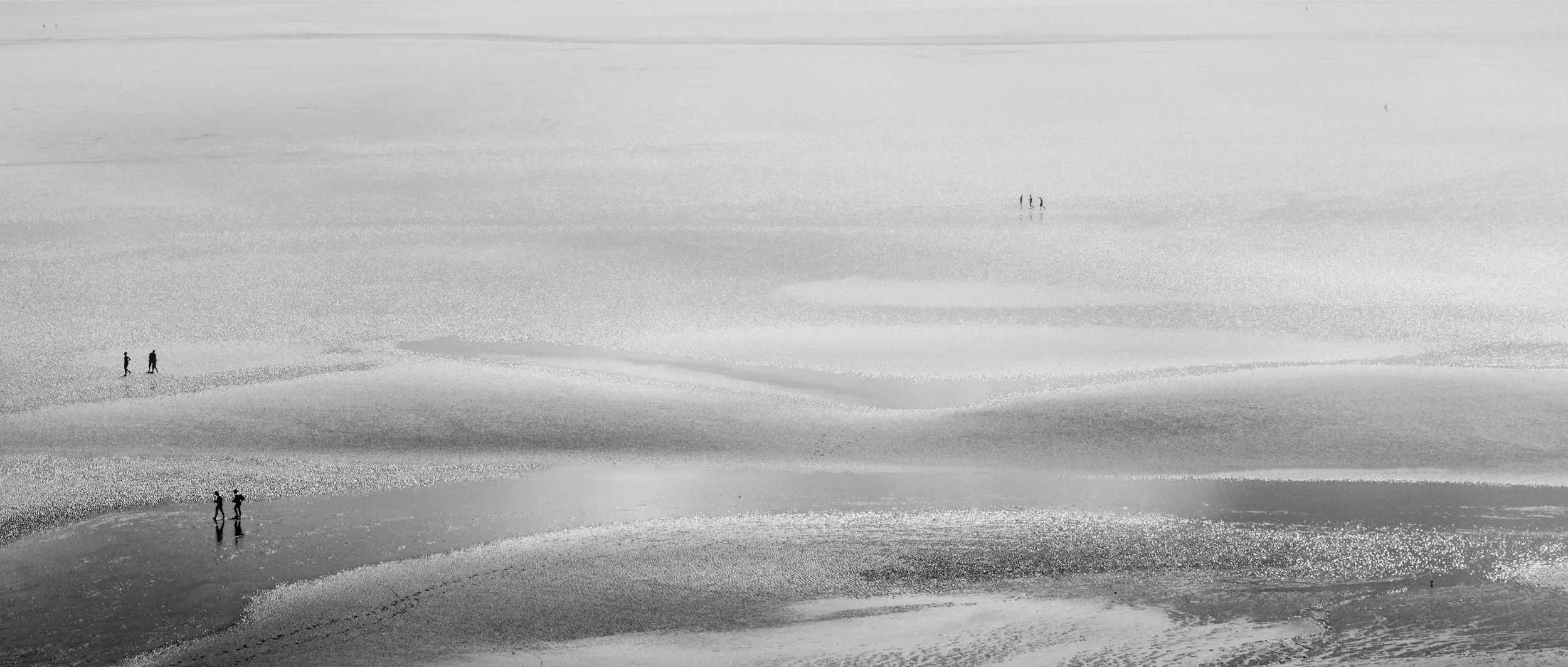 Black and white photo of a vast, shallow body of water with several groups of people walking and standing on the wet sand or shallow areas, creating reflections, with minimal visibility of the horizon.