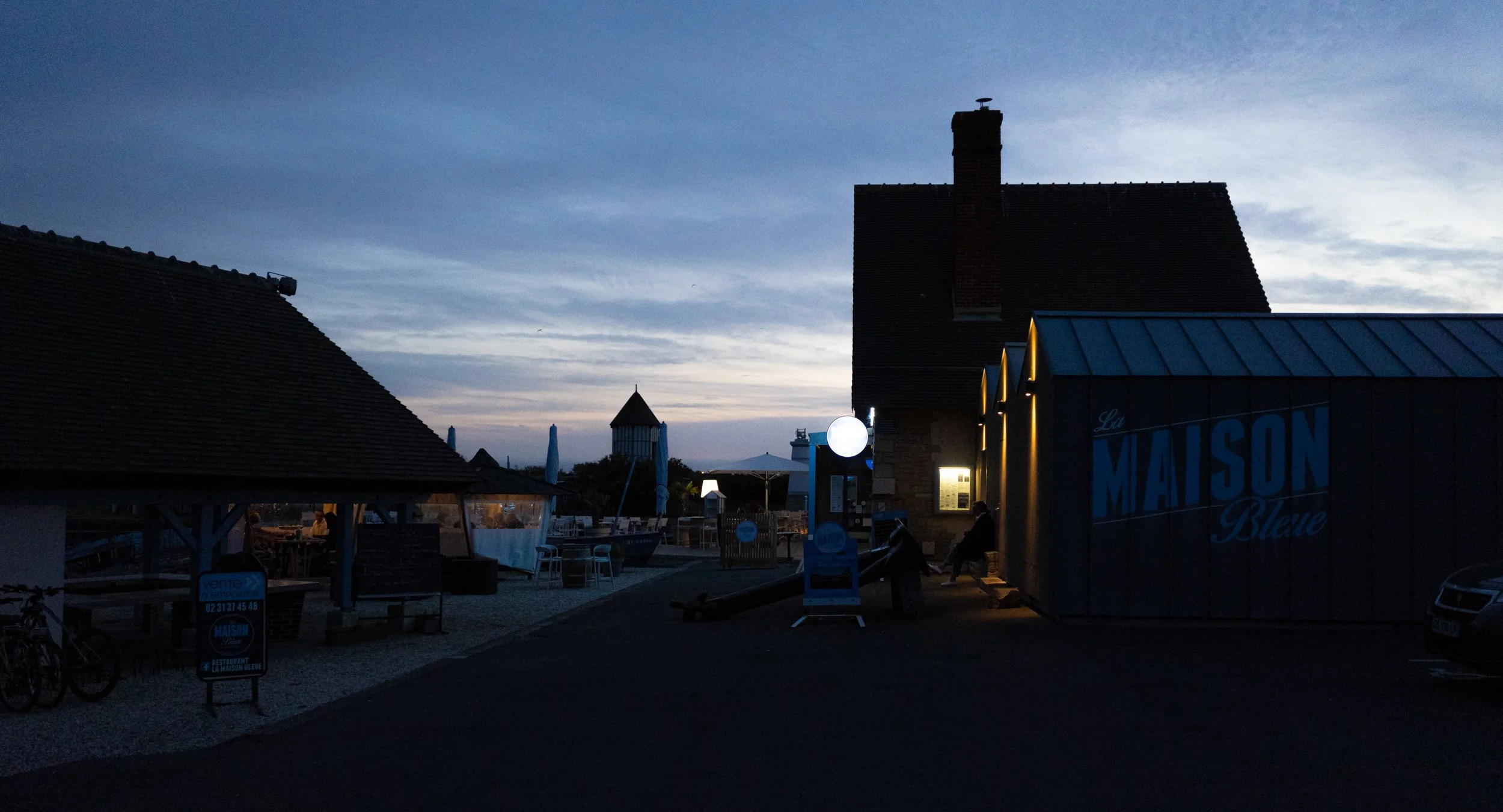 A dimly lit outdoor area during dusk with buildings, umbrellas, and signs, including a sign reading 'La Maison Blue' on a structure. The sky is partly cloudy with a bluish hue, and there are various seating areas and a clock on the building.