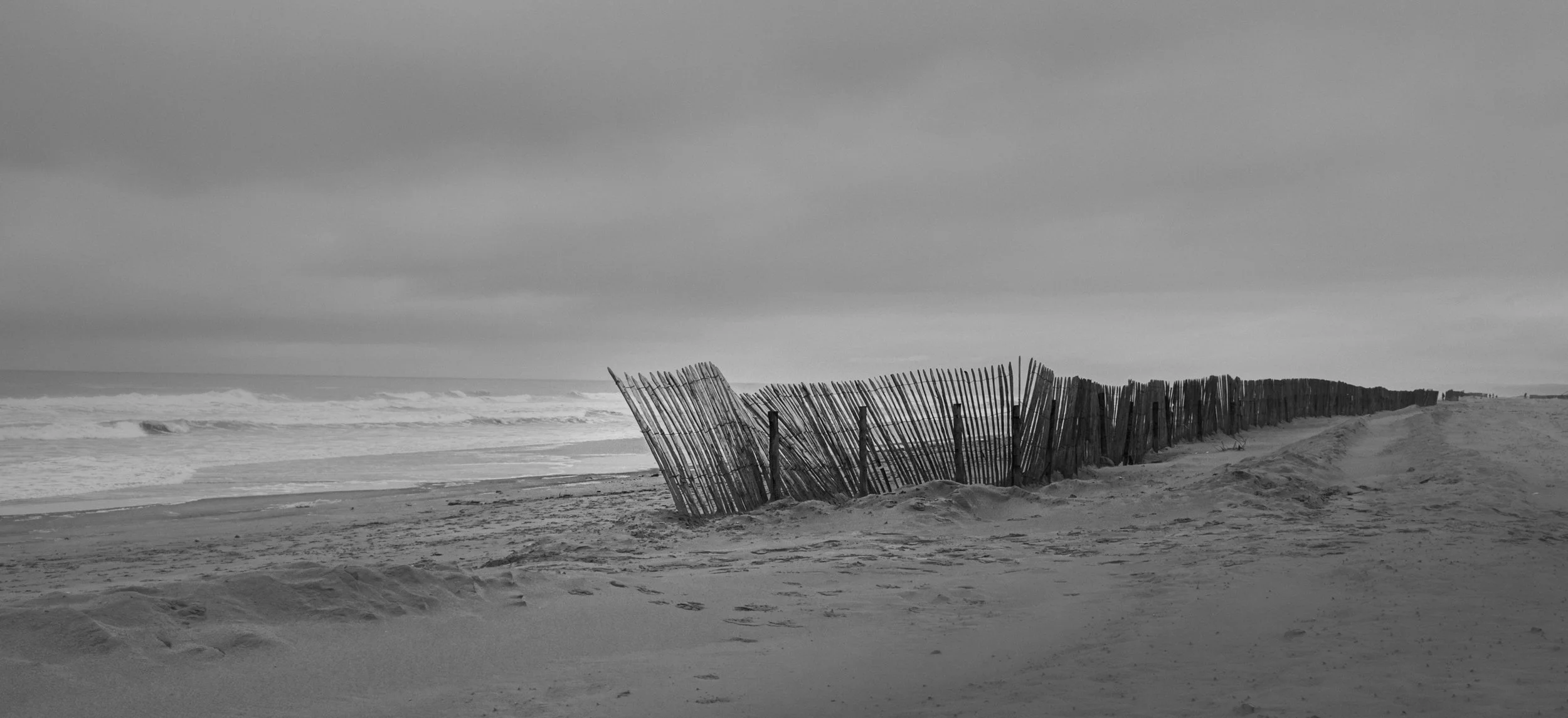 Black and white photo of a beach with a row of worn wooden sand or snow fencing stretching into the distance, with ocean waves and a cloudy sky in the background.