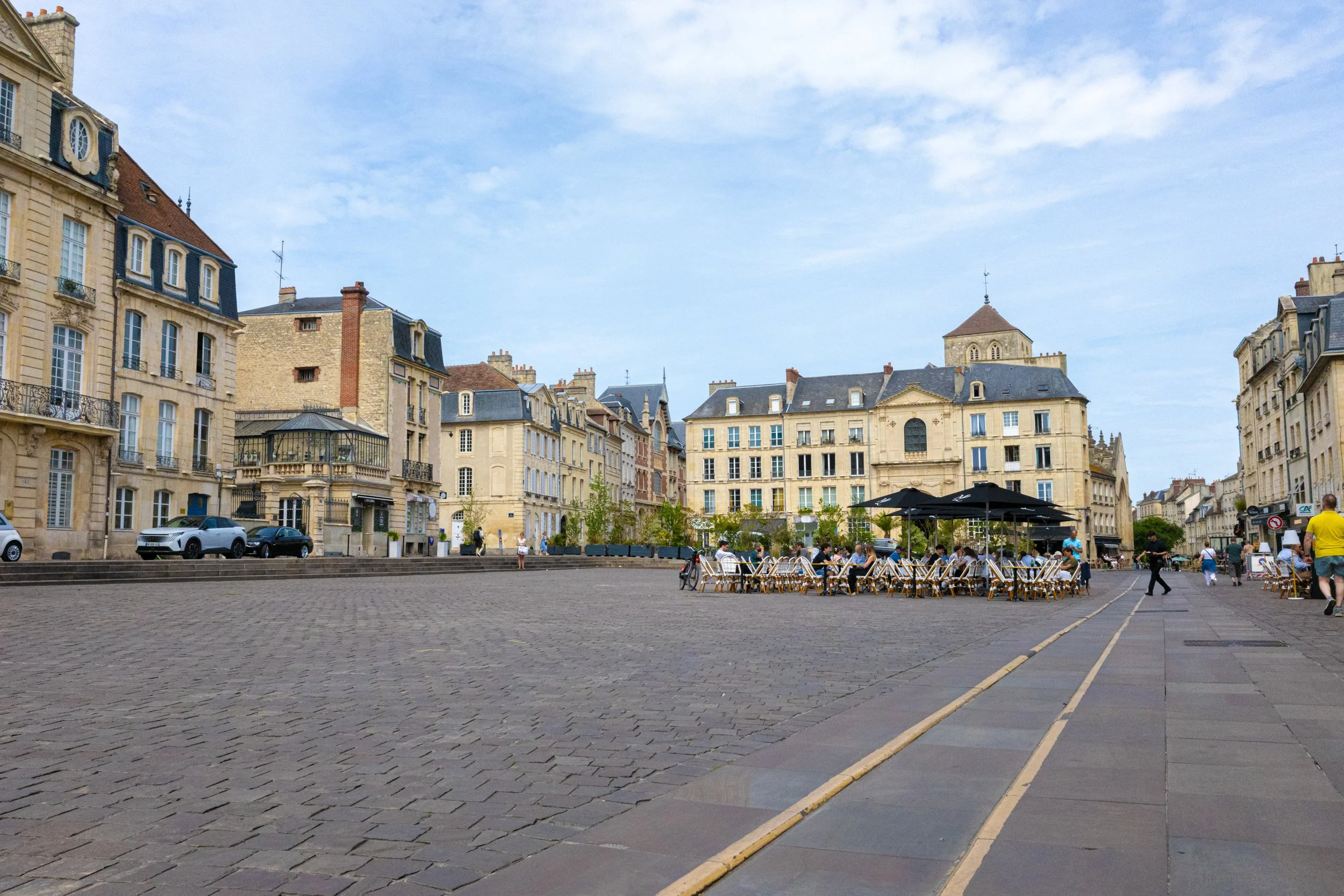 City square with historic European-style buildings, outdoor café seating, people walking and sitting, under a partly cloudy sky.
